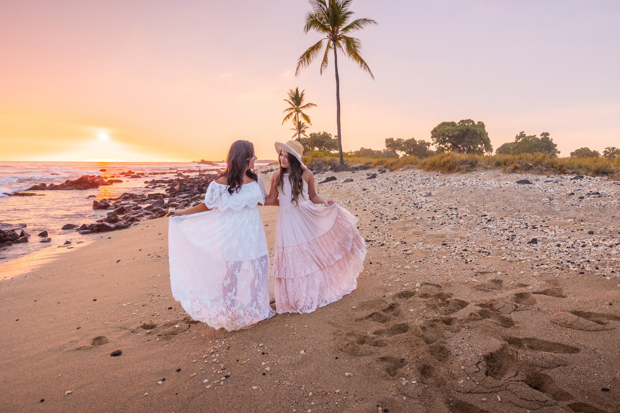 Two women in flowing dresses walk hand in hand on a beach during a sunset, with palm trees and rocks in the background.