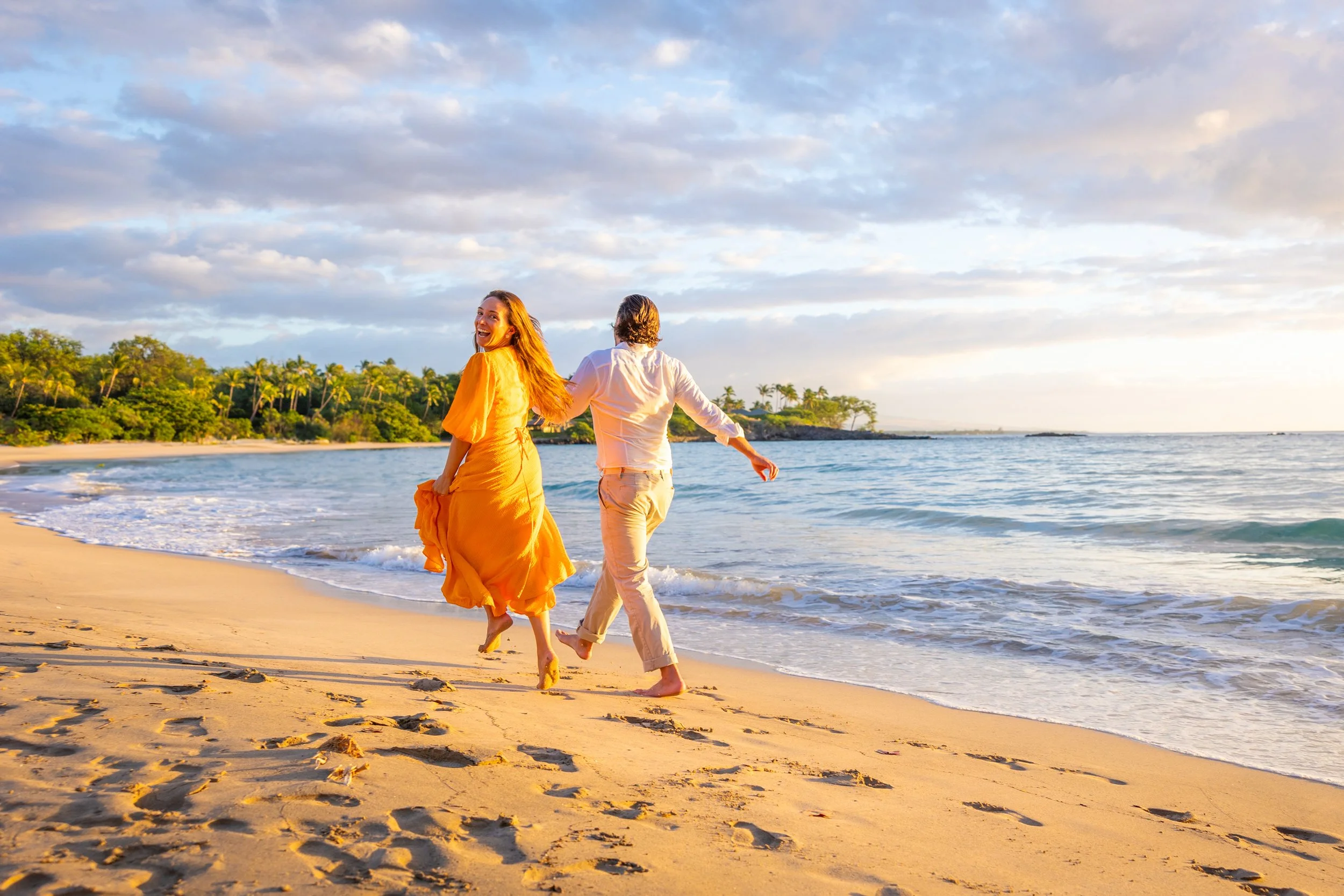 A couple walking hand in hand along the beach at sunset, with the woman wearing a yellow dress and the man in white clothes, near the shoreline with sand and ocean waves, and a tropical forest in the background.