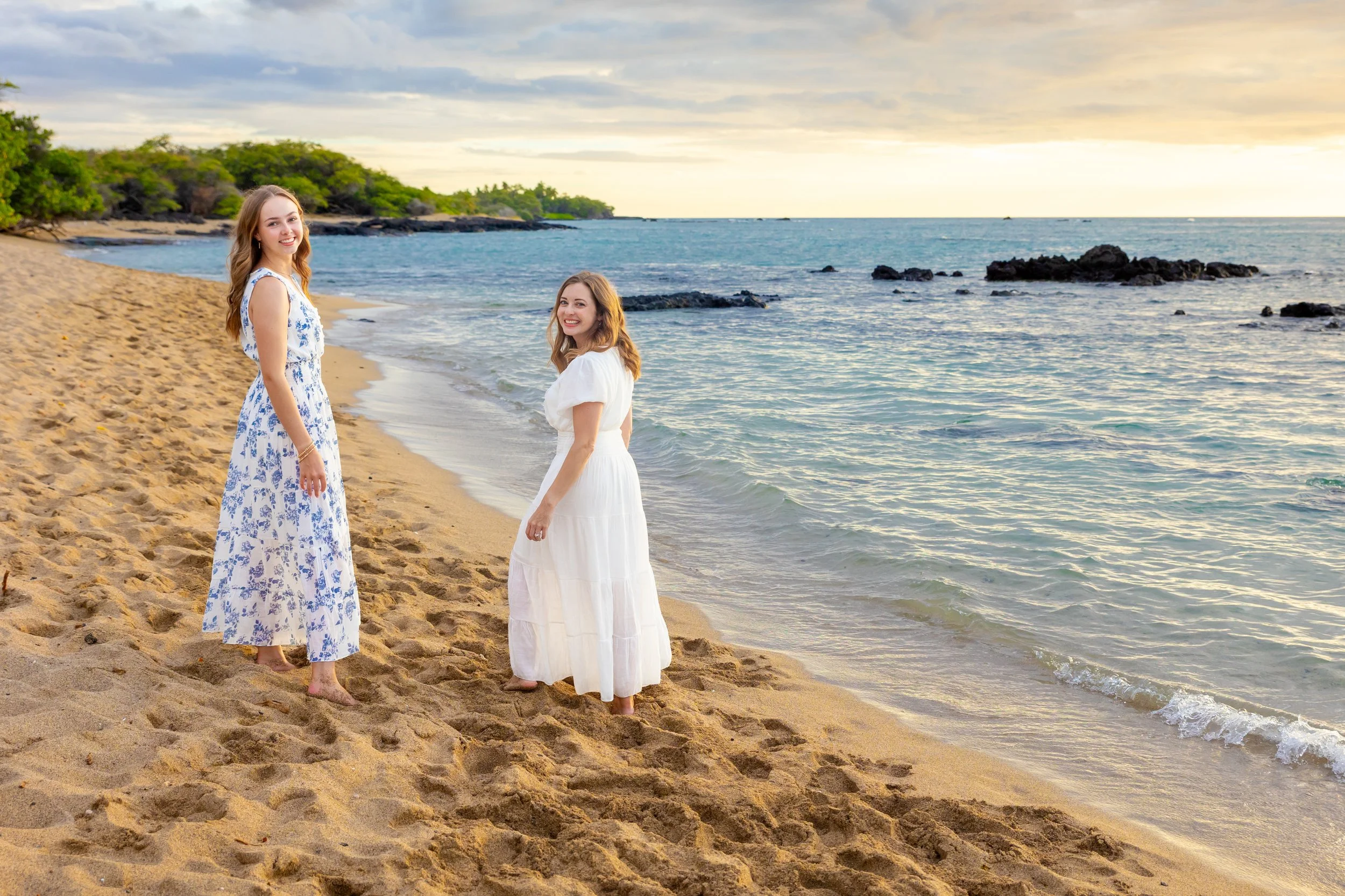 Two women in white dresses standing on a sandy beach near the water, with trees and rocks in the background, during sunset.