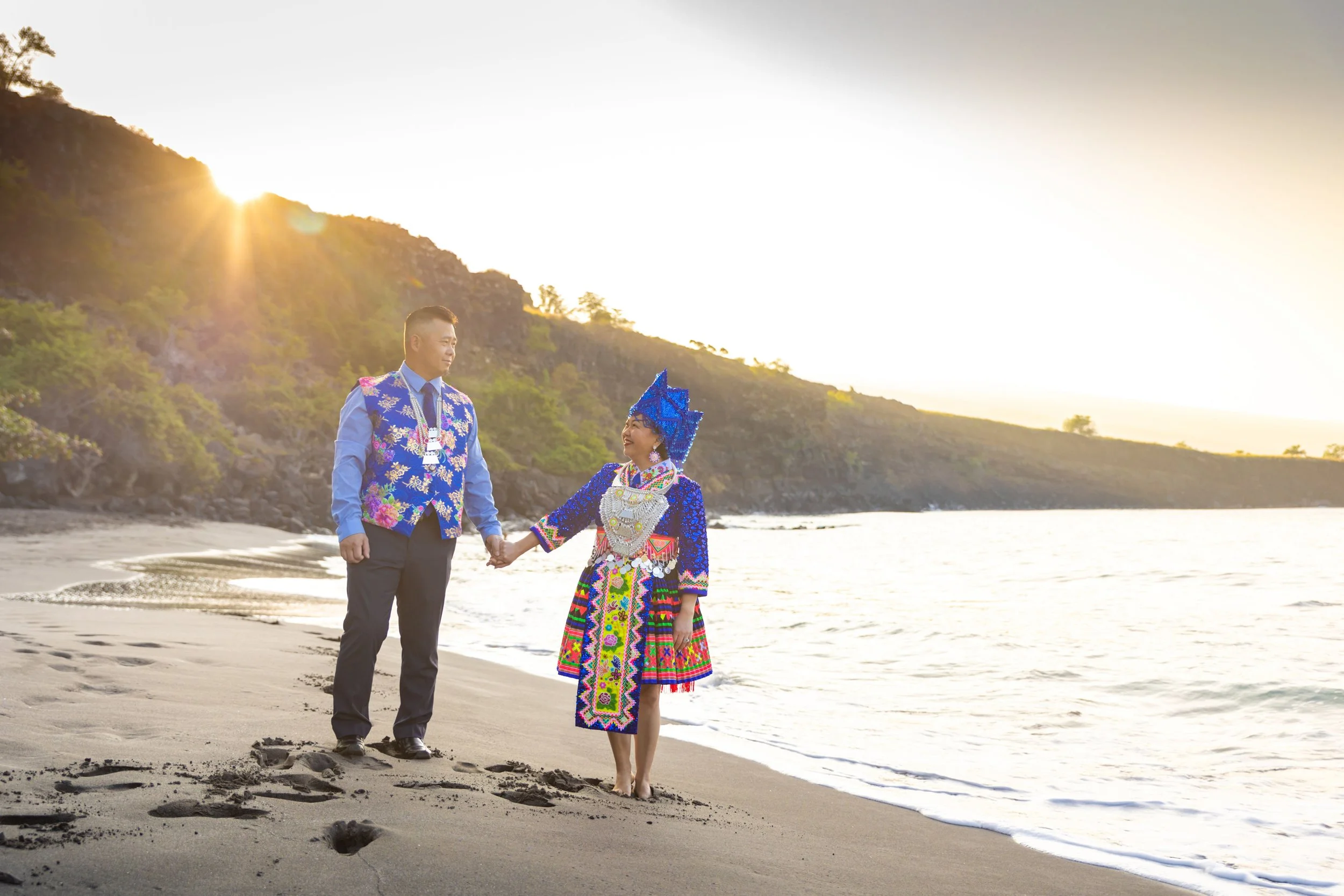 A couple holding hands on a beach at sunset, with the man wearing a colorful traditional outfit and the woman dressed in vibrant, ethnic clothing with a tall headpiece.