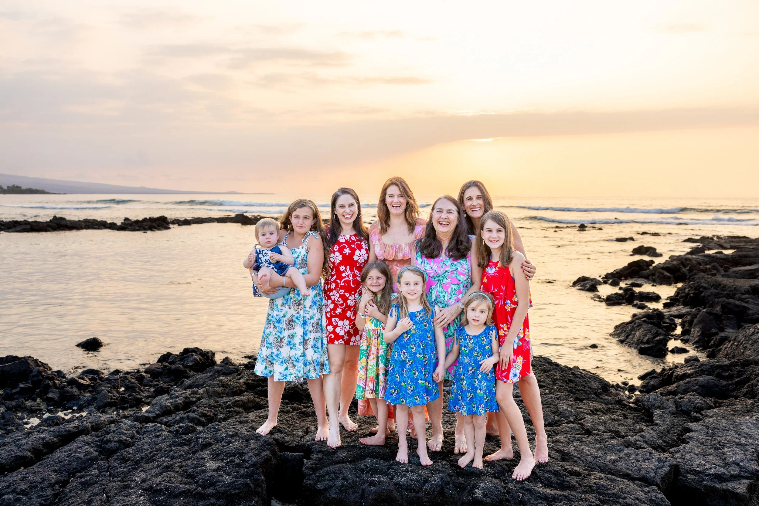 A large group of women and children standing on rocks at the beach during sunset, all wearing colorful floral dresses, smiling and posing for the photo.