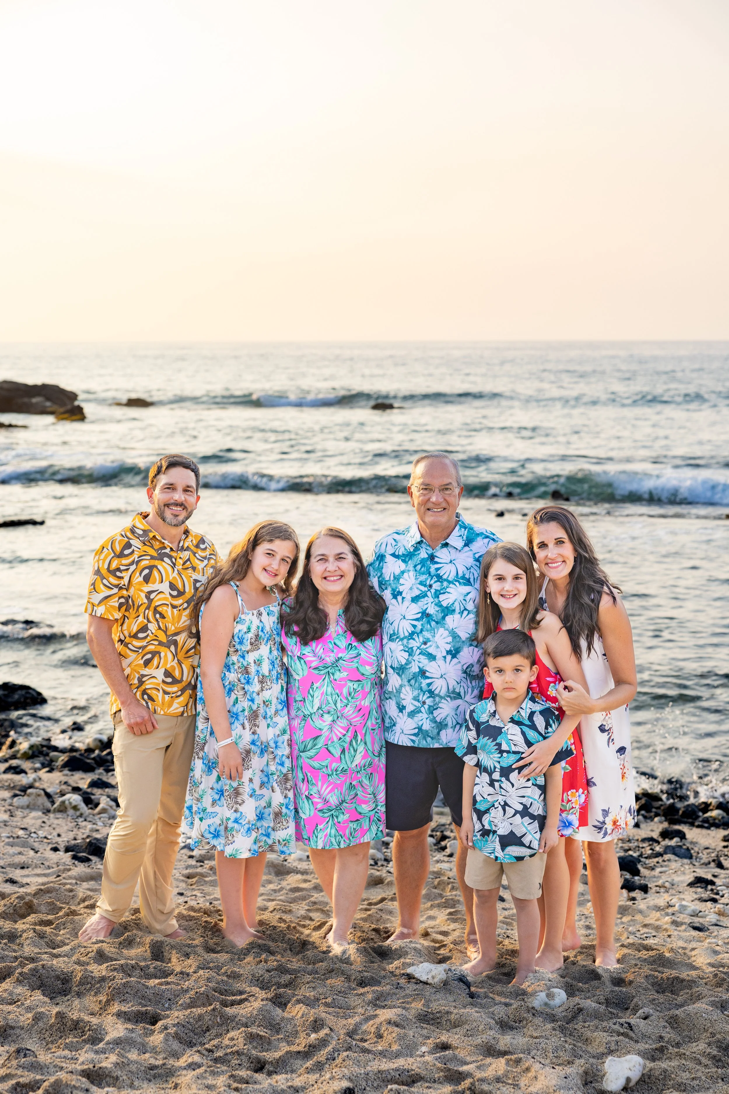 Family of eight posing on a beach at sunset, dressed in colorful summer outfits.