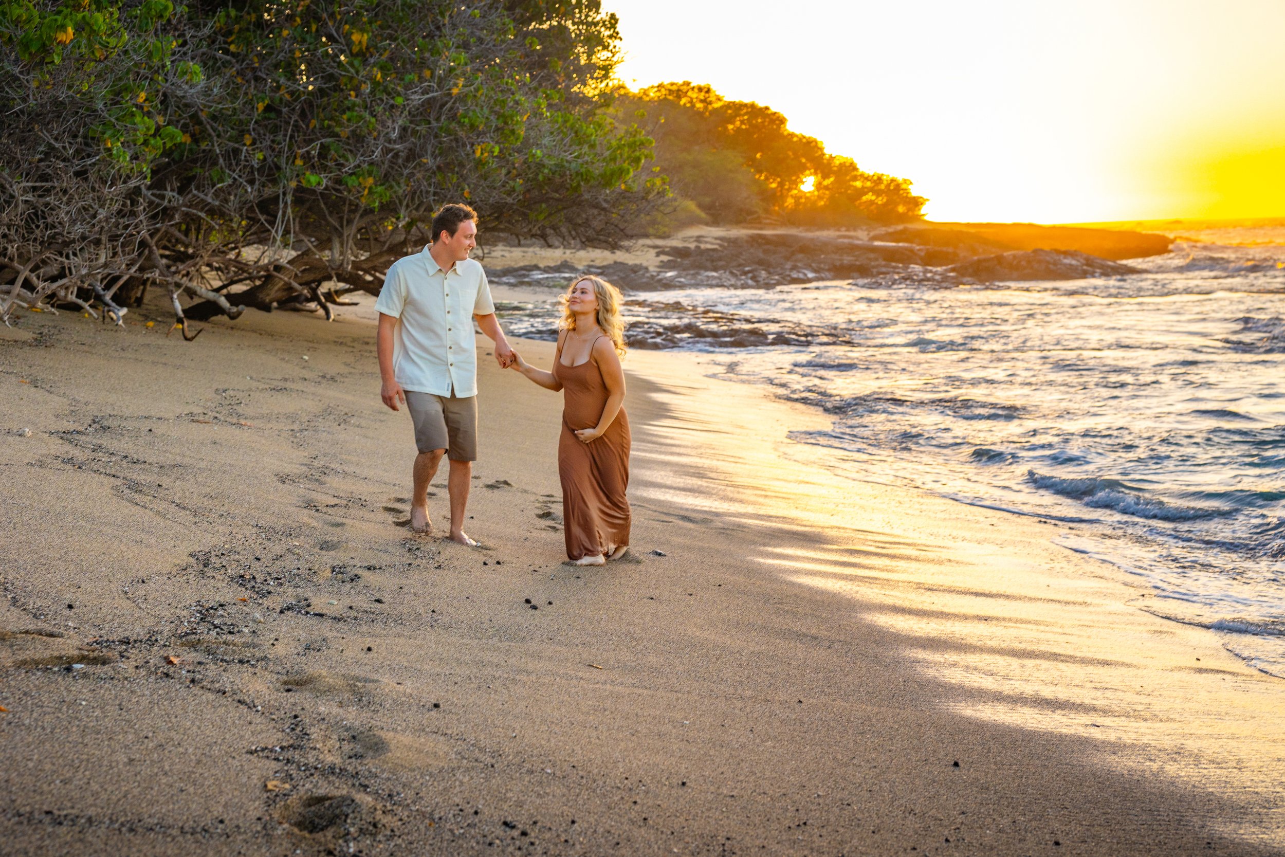 A man and woman walking hand in hand on a beach at sunset, with trees on the left and the ocean waves on the right.