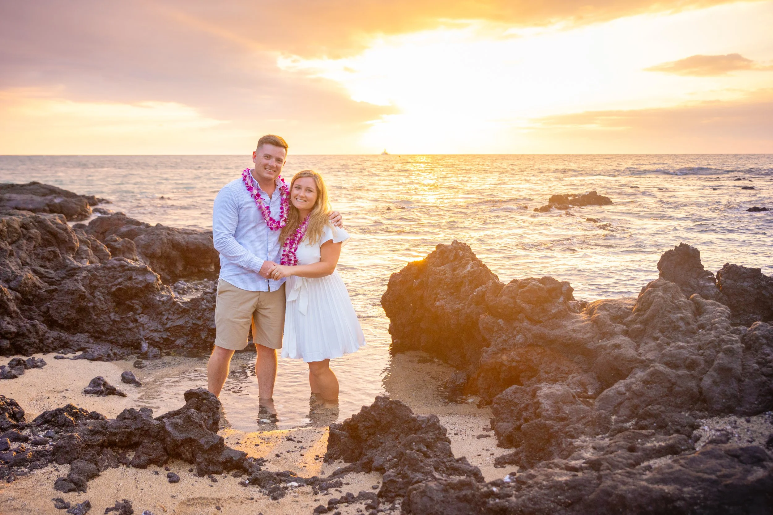 Couple standing in the water on a rocky beach at sunset, wearing leis, smiling at the camera.