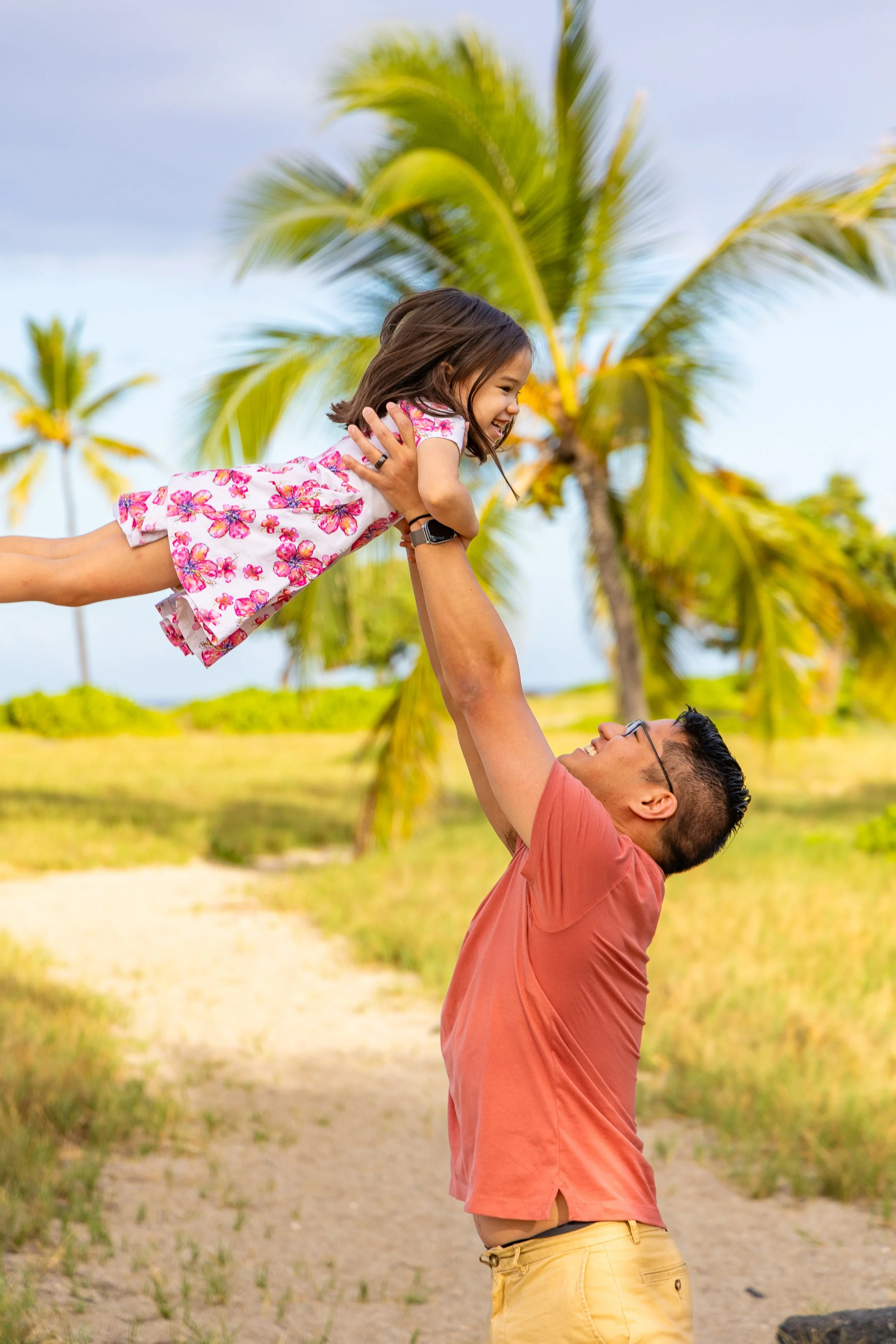 A man lifting a young girl in a pink floral dress into the air on a sandy path surrounded by palm trees.