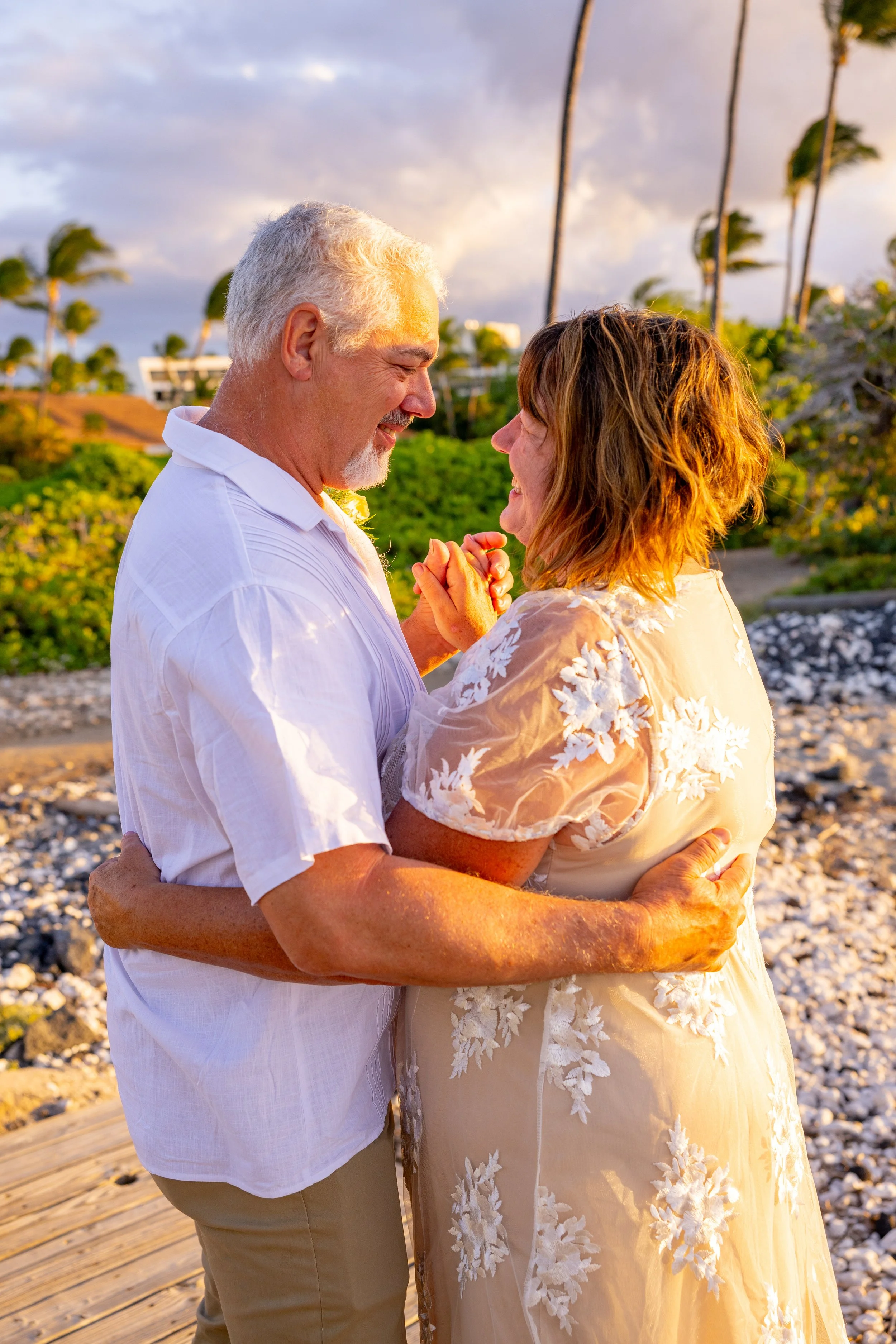 A senior couple dancing and smiling outdoors on a beach with palm trees and greenery during sunset.