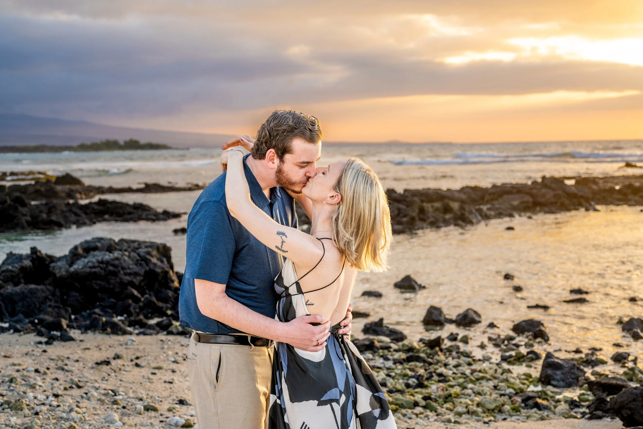 A couple sharing a kiss on the beach during sunset, with the ocean and rocky shoreline in the background.
