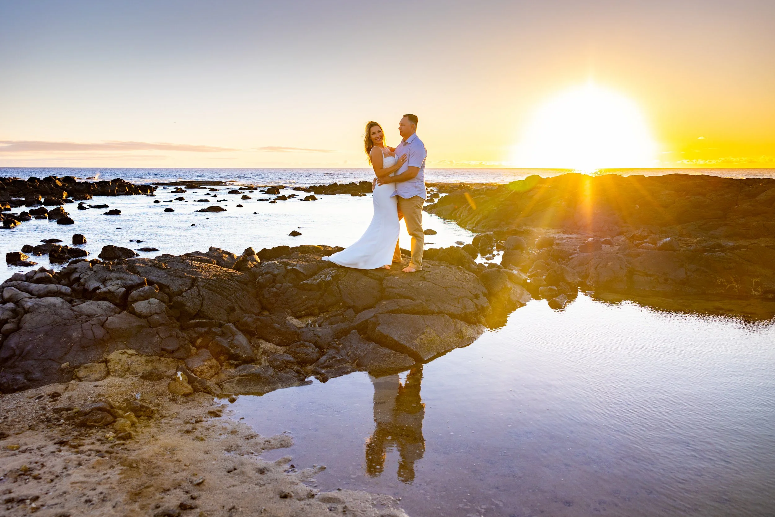 A couple in wedding attire standing on rocks by the ocean at sunset, with the bride in a white dress and the groom in a light shirt and beige pants, embracing each other.