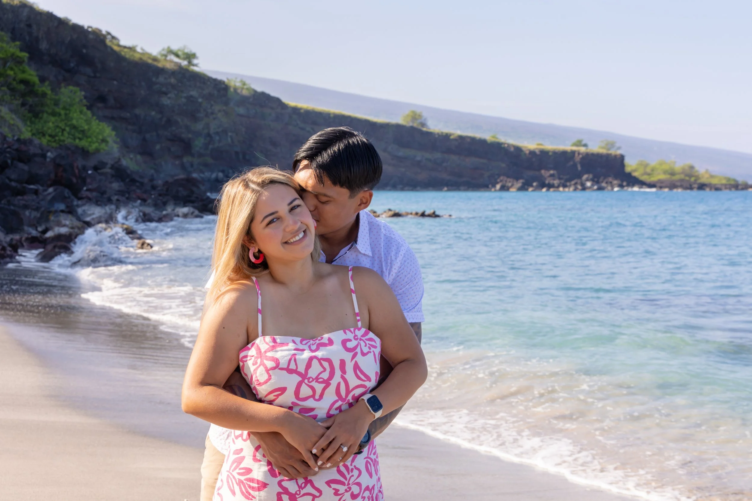 A couple standing on a beach with ocean and cliffs in the background. The woman is smiling, wearing a pink and white floral dress, and the man is kissing her on the cheek.