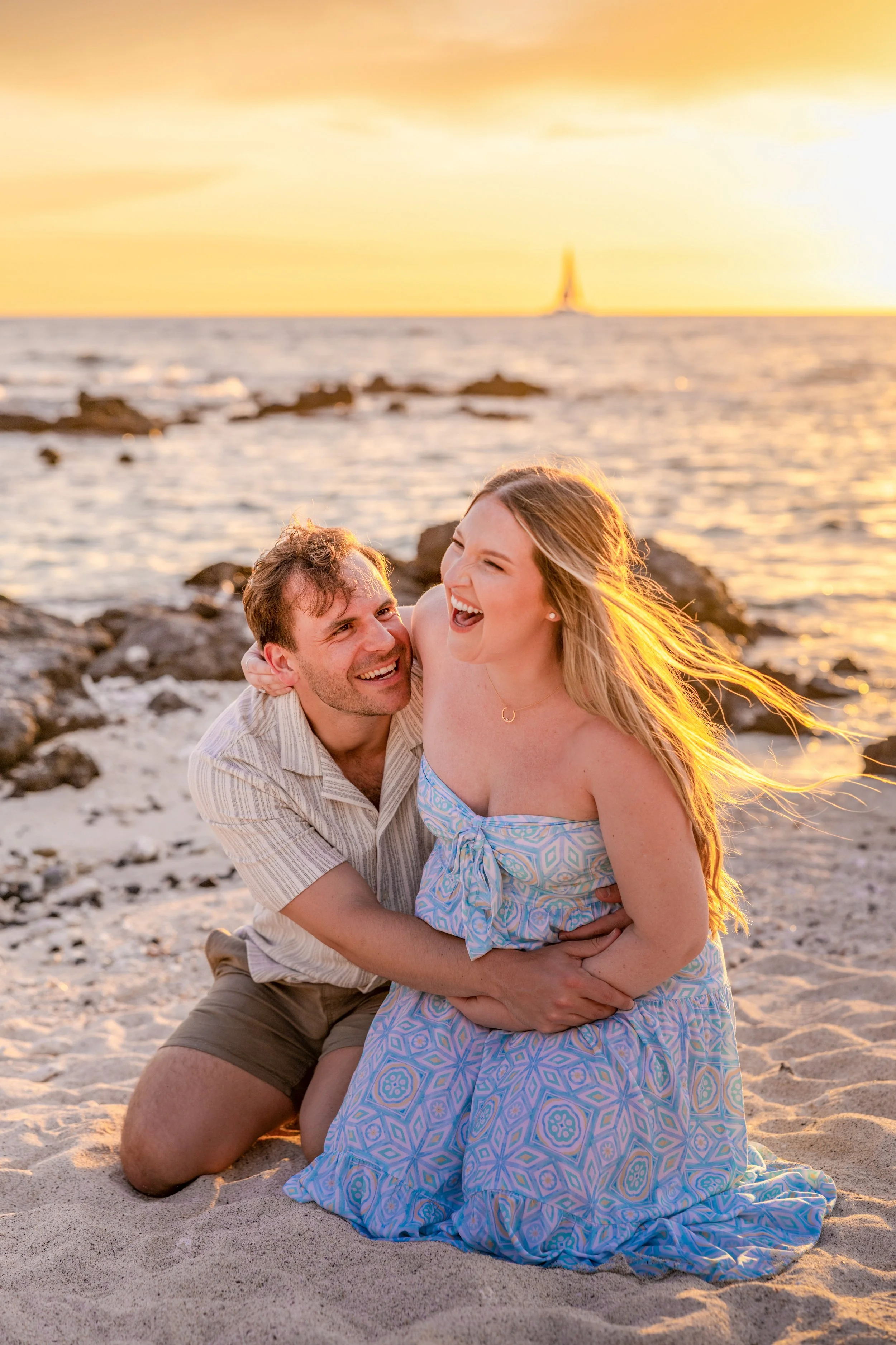 A couple is laughing and hugging on a beach at sunset, with the ocean and a lighthouse in the background.