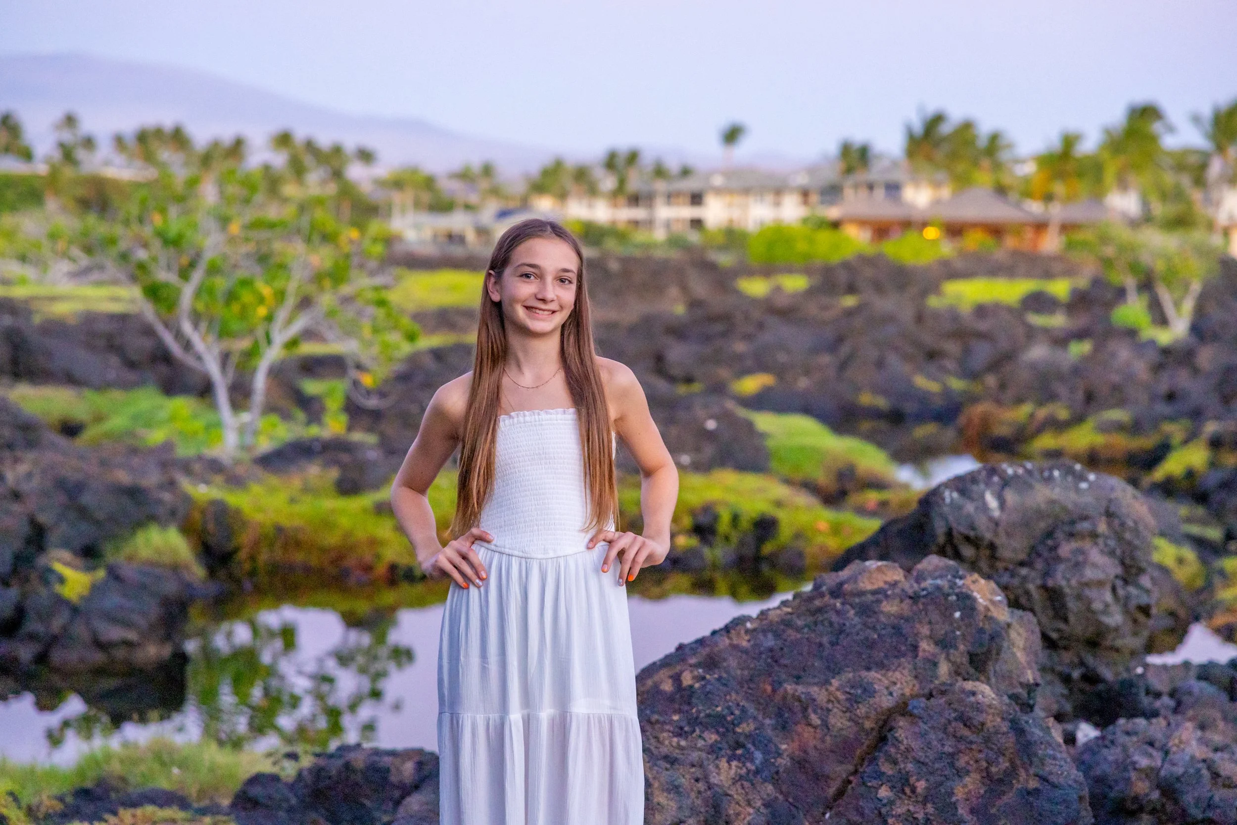 A young girl with long brown hair, wearing a white strapless dress, standing outdoors on rocky terrain with water, trees, houses, and mountains in the background during sunset.
