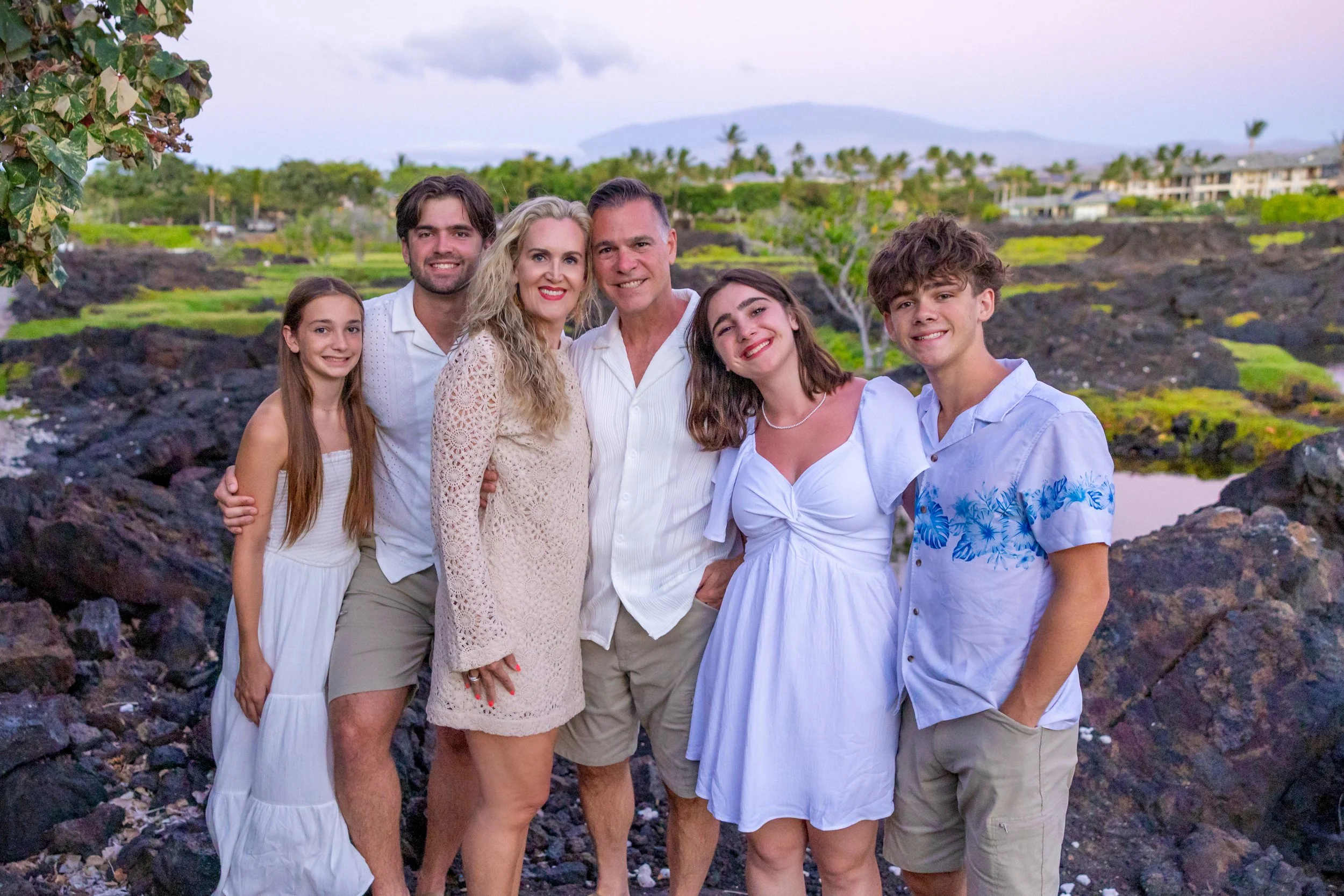 A family of seven standing outdoors on a rocky landscape with lush greenery and distant hills in the background, all dressed in light-colored summer clothing, smiling at the camera.