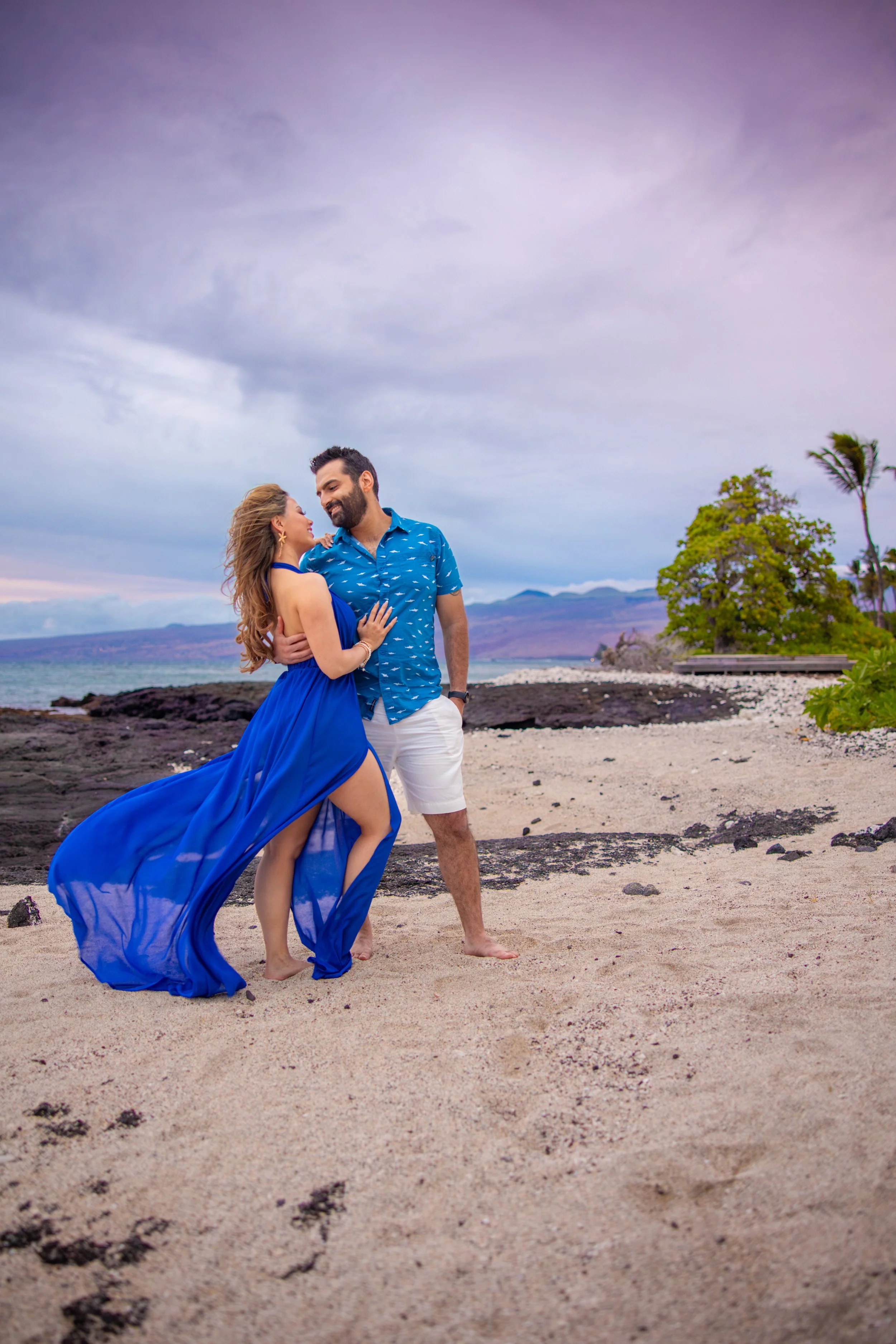 A couple dressed in blue holding each other on a beach with black rocks, trees, and mountains in the background under a cloudy sky.