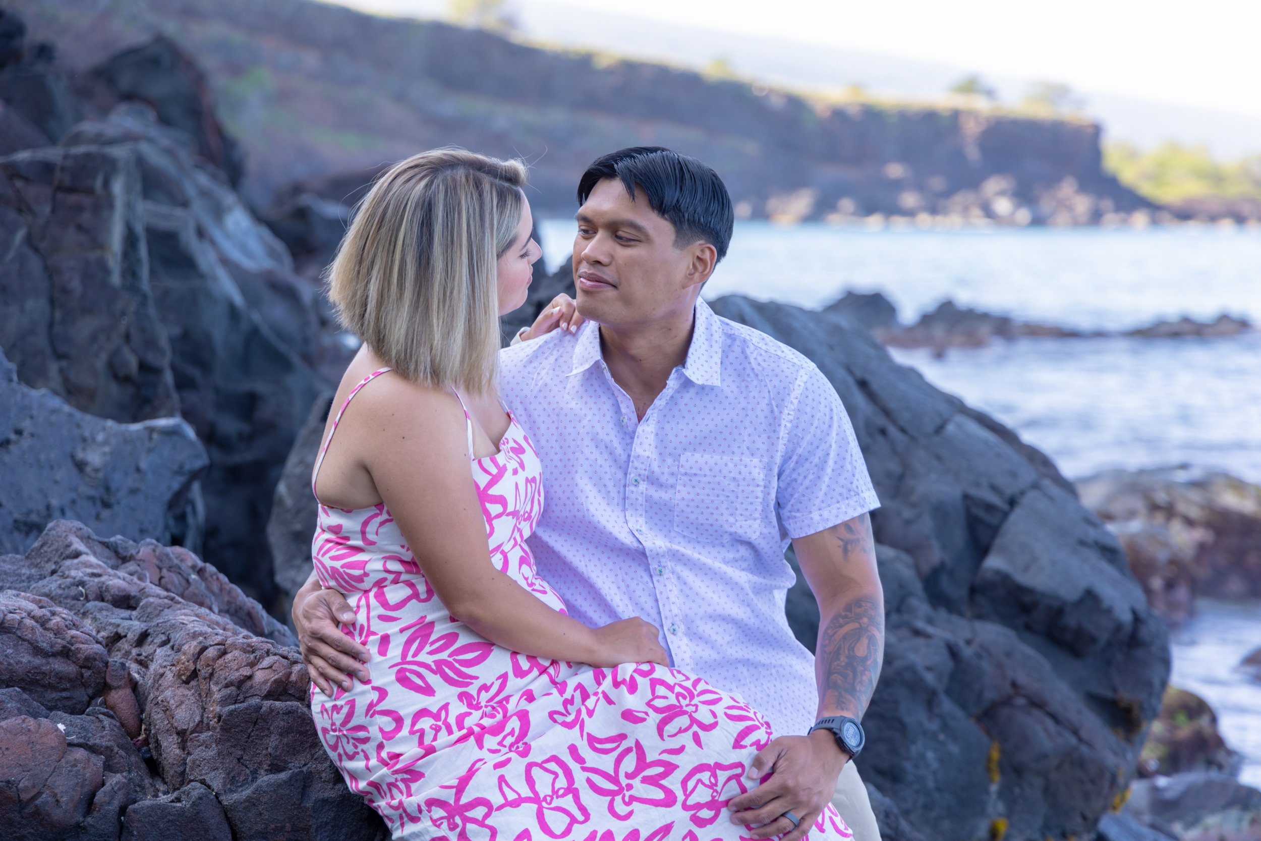 A man and woman sitting close together on rocks by the ocean, gazing at each other with a scenic coastal background.