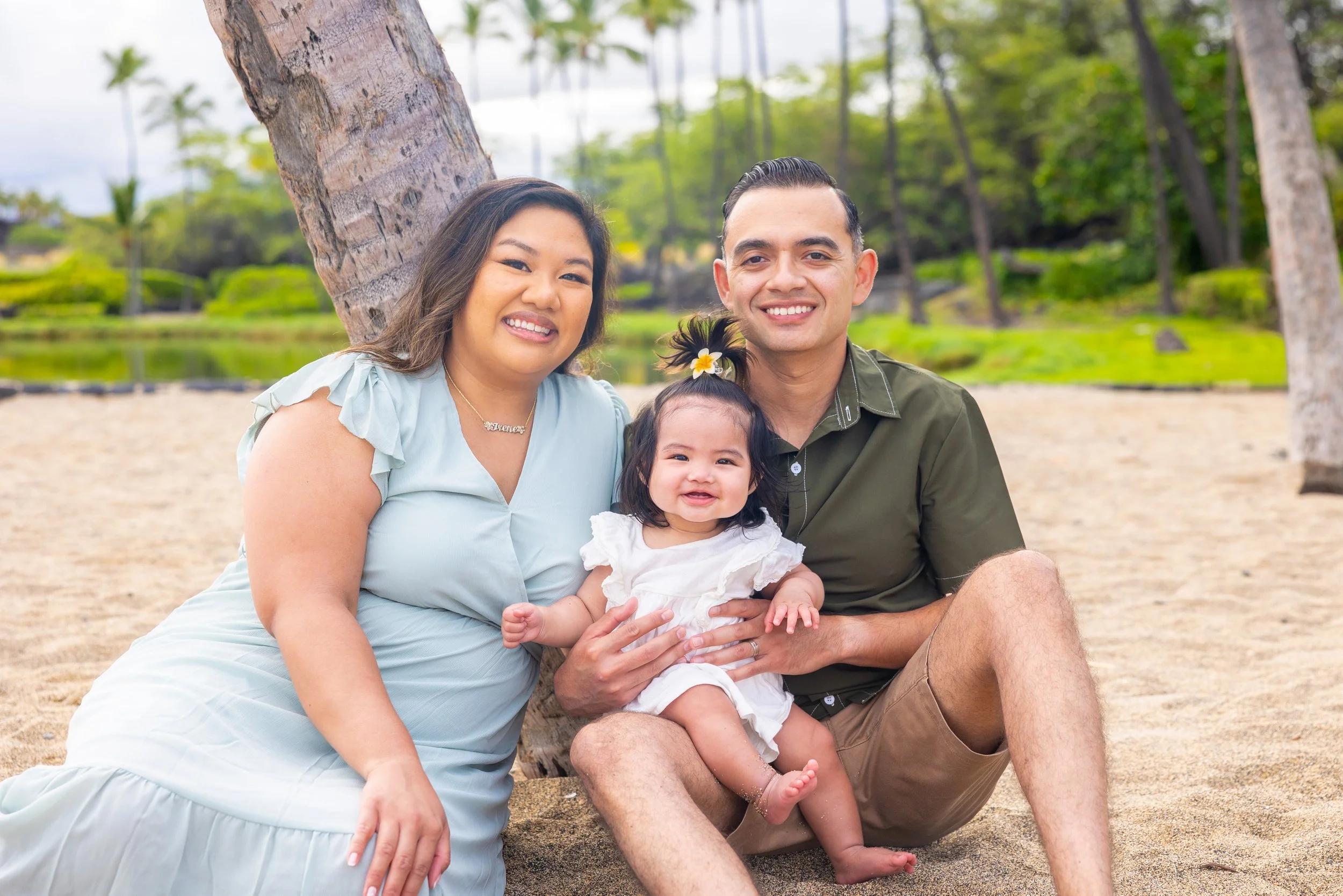 A family of three sitting on the sand near a tree in a park. The woman is wearing a light blue dress, the man is in a green shirt and shorts, and the little girl is in a white dress with a flower in her hair, all smiling at the camera.