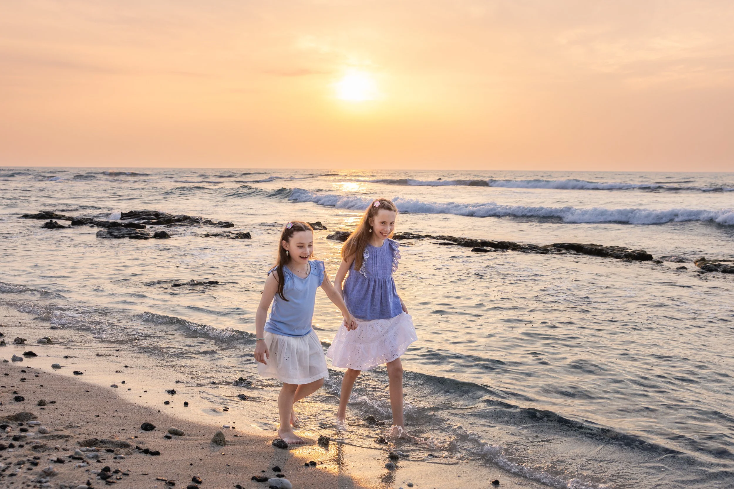 Two young girls in blue and white dresses holding hands and walking in the shallow ocean water at sunset on a sandy beach.