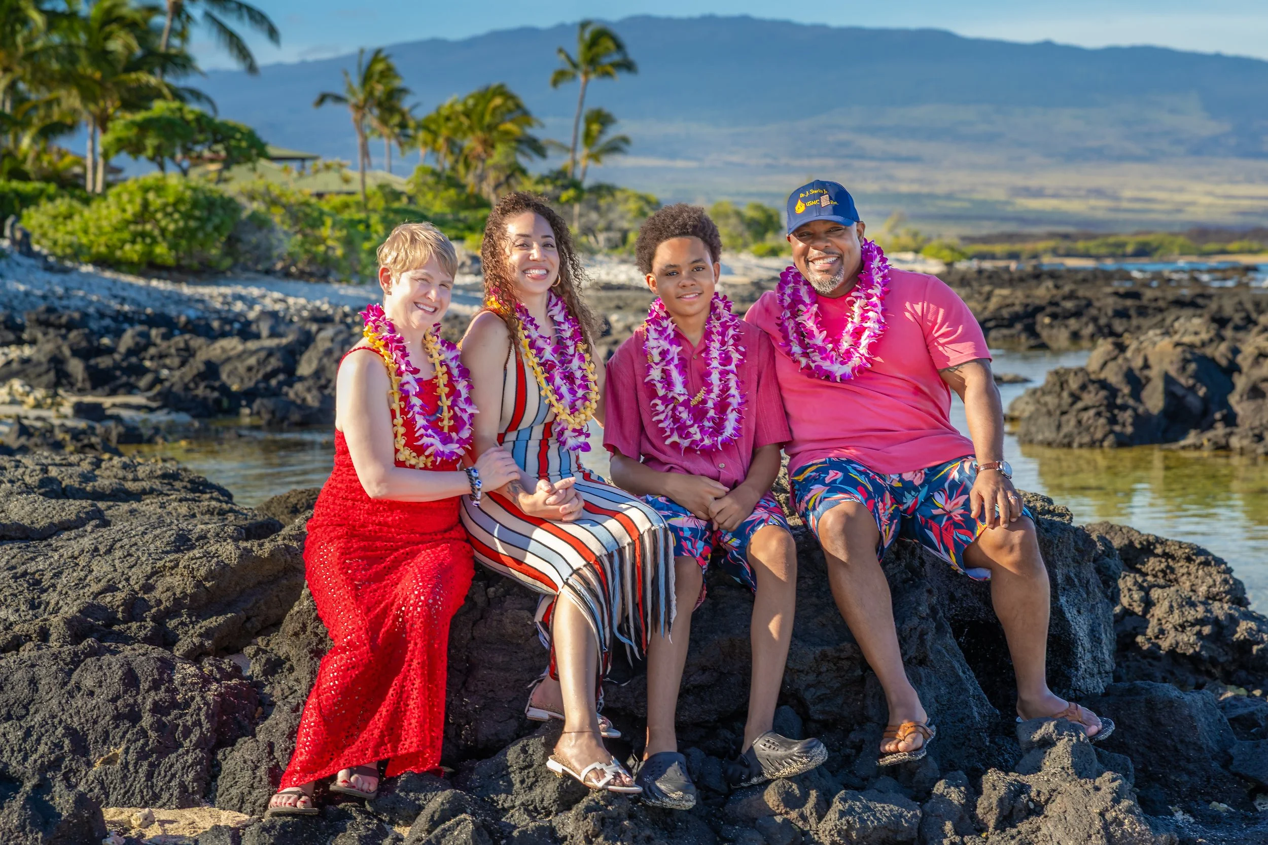 A family of four sitting on rocks by the ocean, wearing Hawaiian leis and casual summer clothing, with palm trees, volcanic rocks, and mountains in the background.
