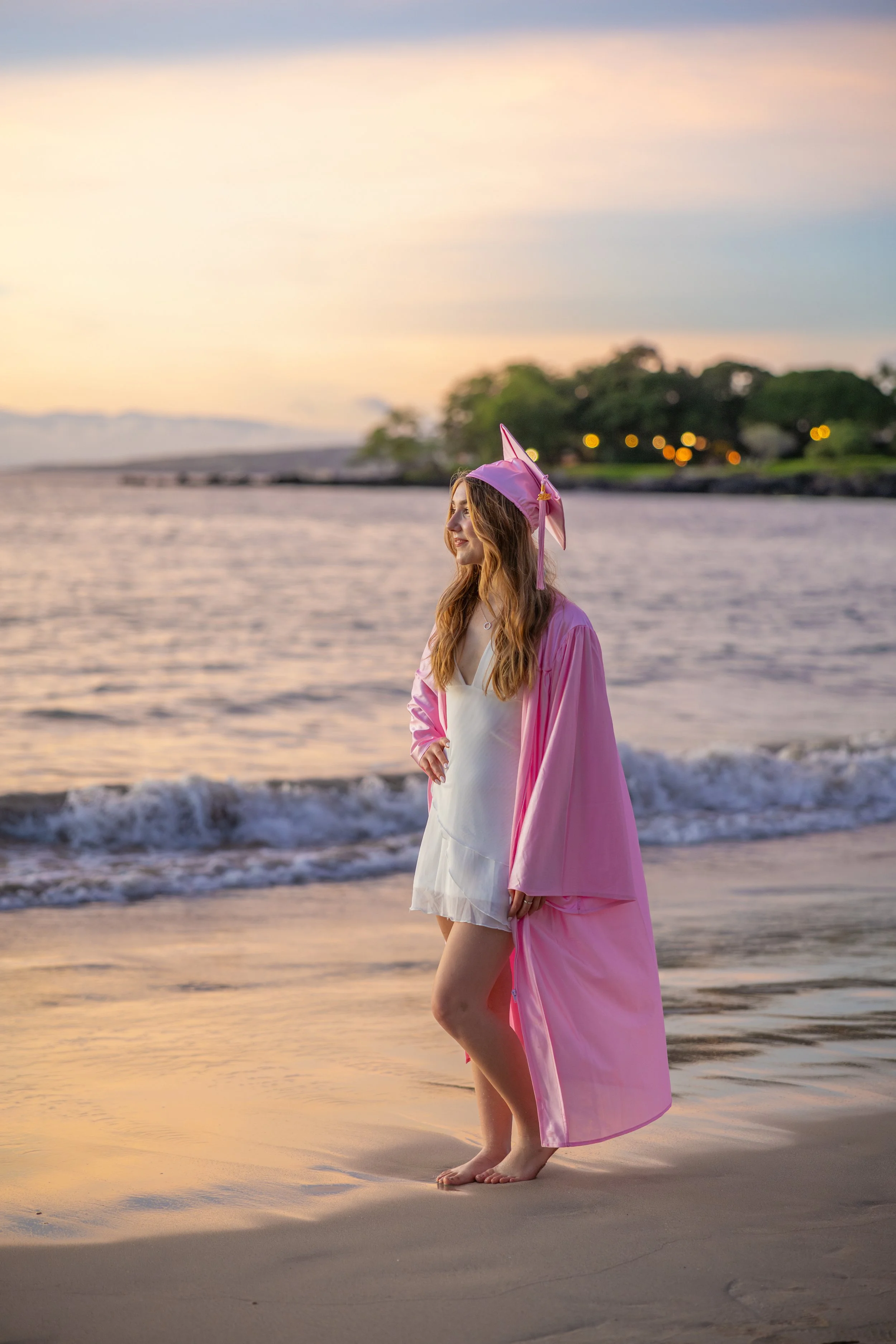 A woman standing barefoot on a beach during sunset, wearing a pink graduation gown and a pink graduation cap with a unicorn horn, looking to the side with a smile.