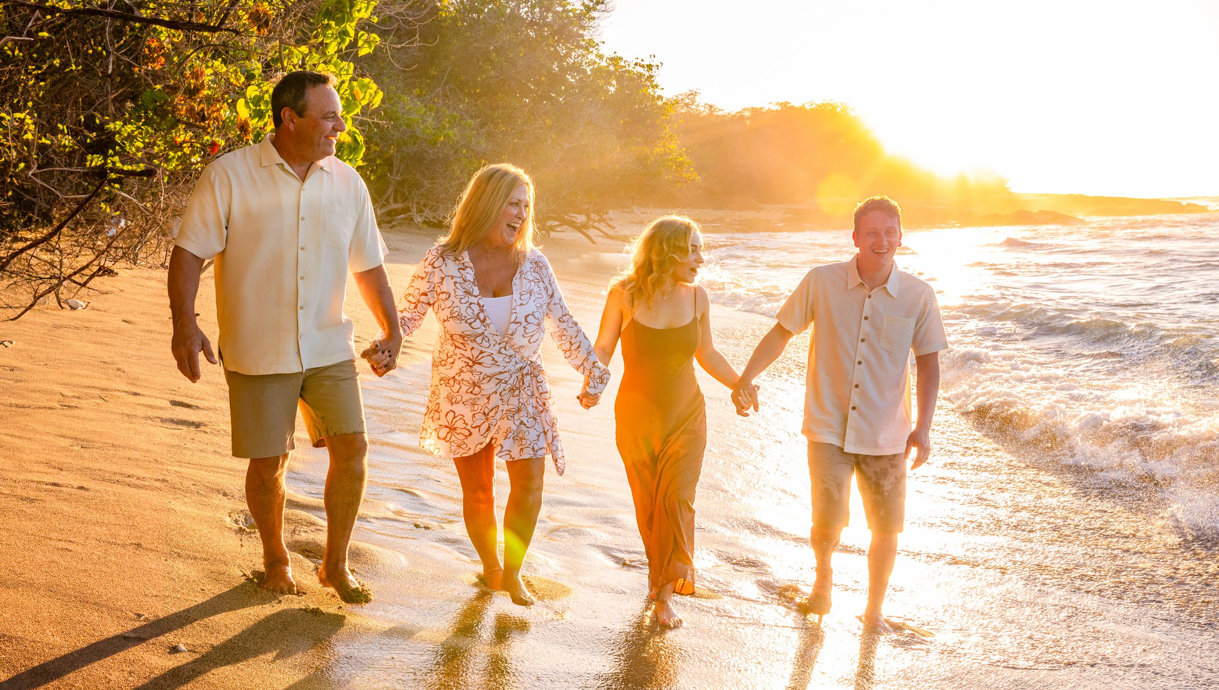 A family of five walking hand in hand along a beach at sunset, with waves and trees in the background.