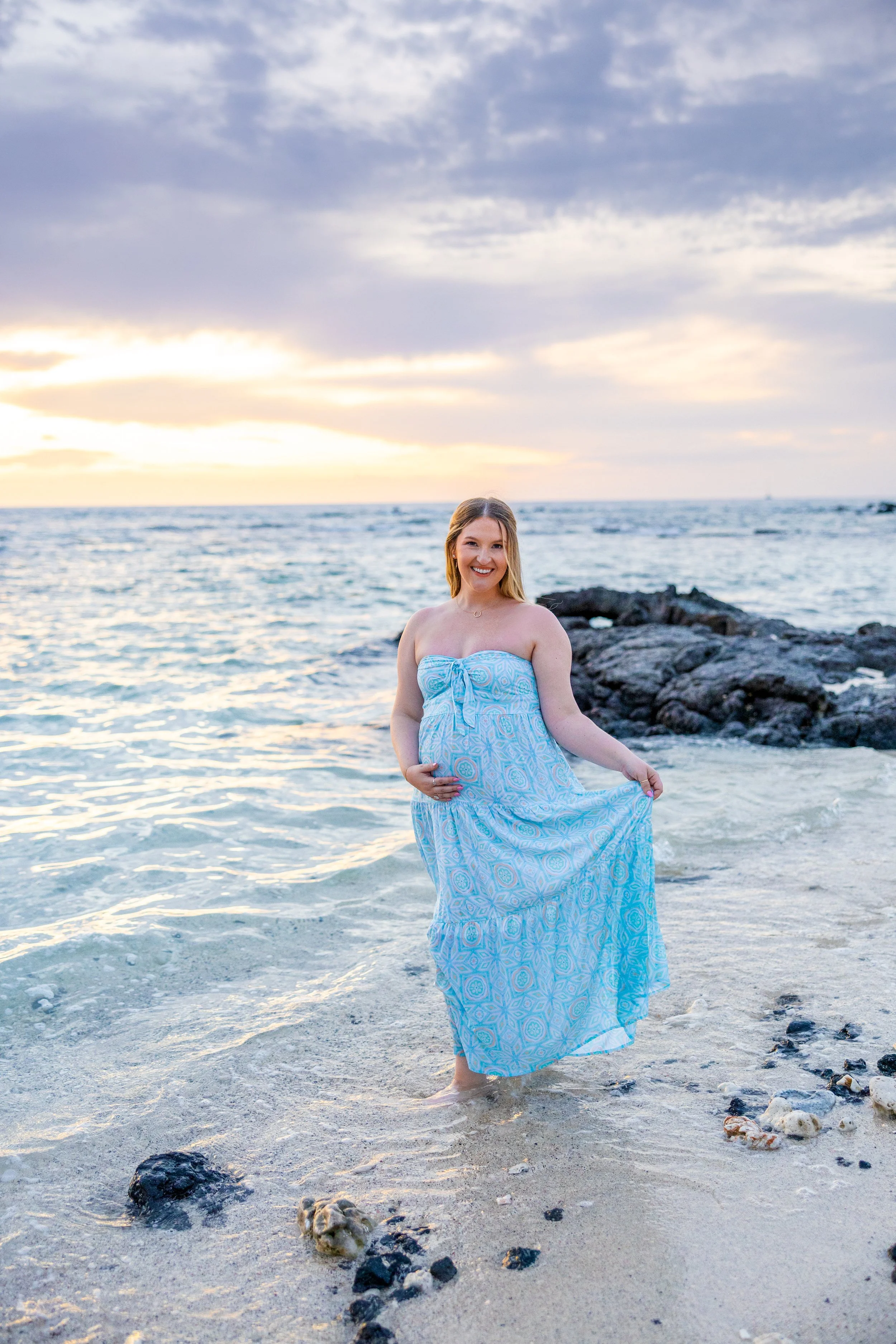A pregnant woman wearing a blue patterned dress standing on the beach with the ocean and rocks in the background at sunset.