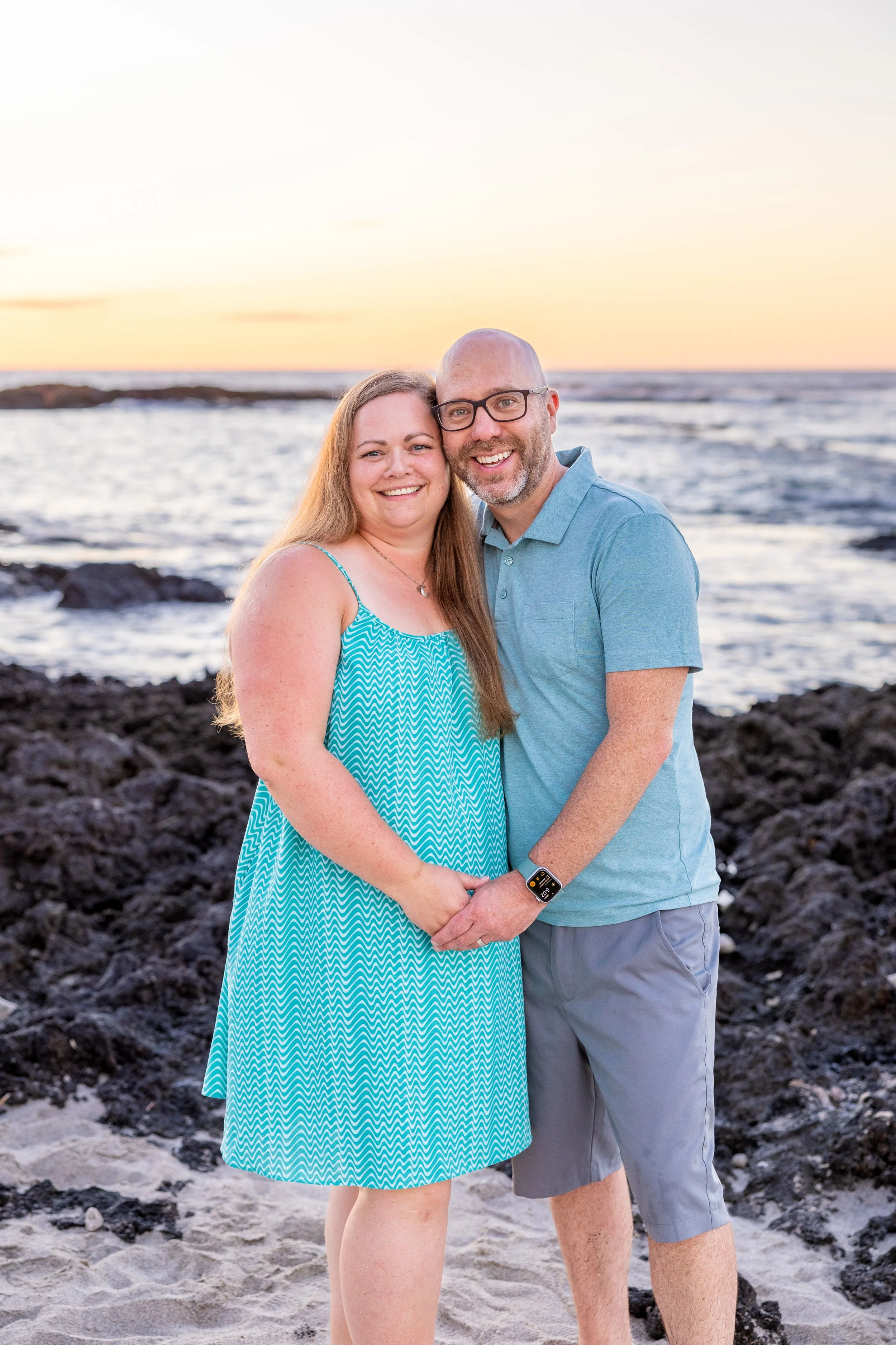 A smiling couple standing hand in hand on a beach with rocky shoreline at sunset, ocean waves in background.