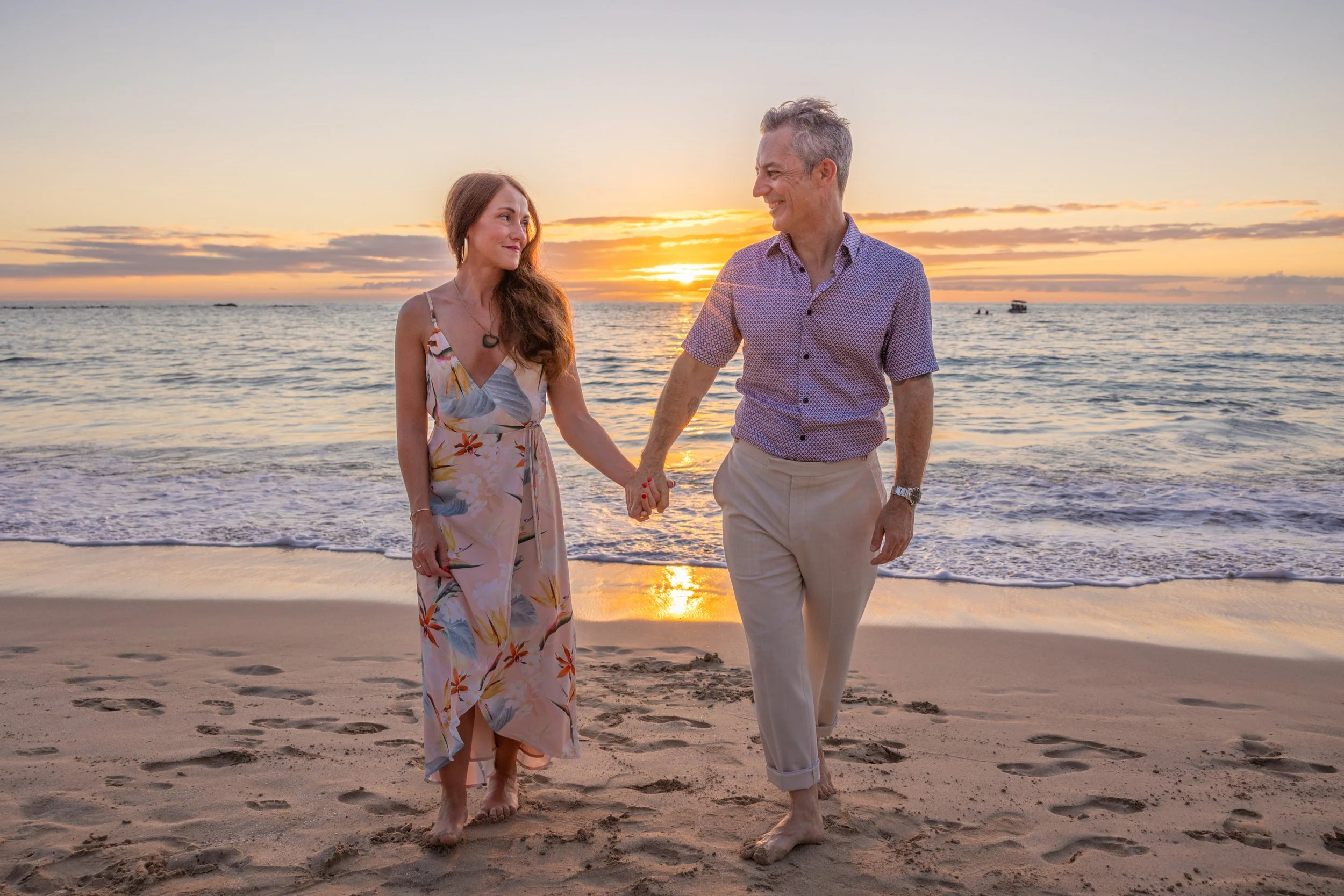 A couple holding hands walking on the beach at sunset, with a calm ocean and a few boats in the background.