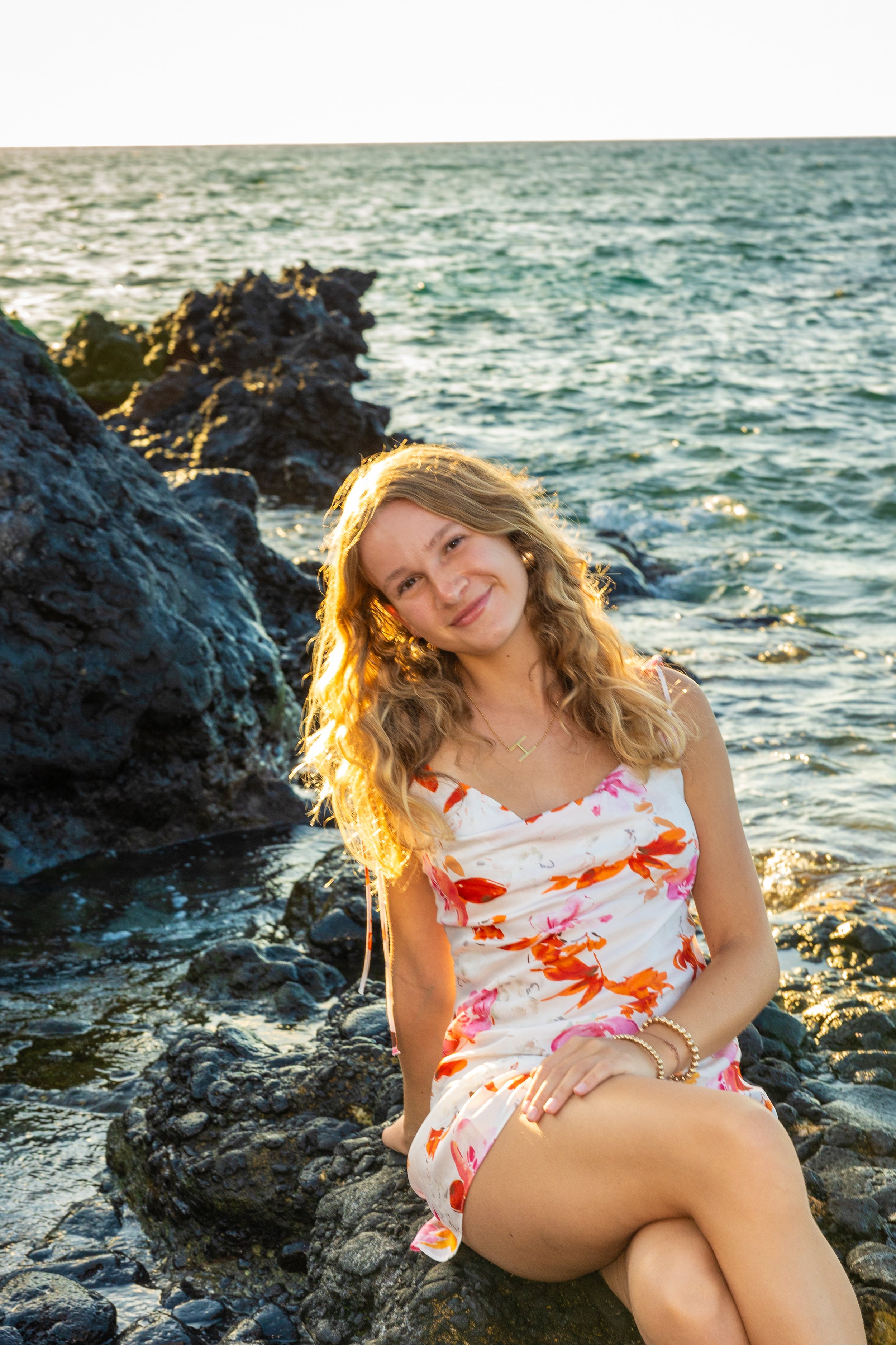 A young woman with long blonde hair sitting on rocks by the ocean, smiling at the camera during sunset.