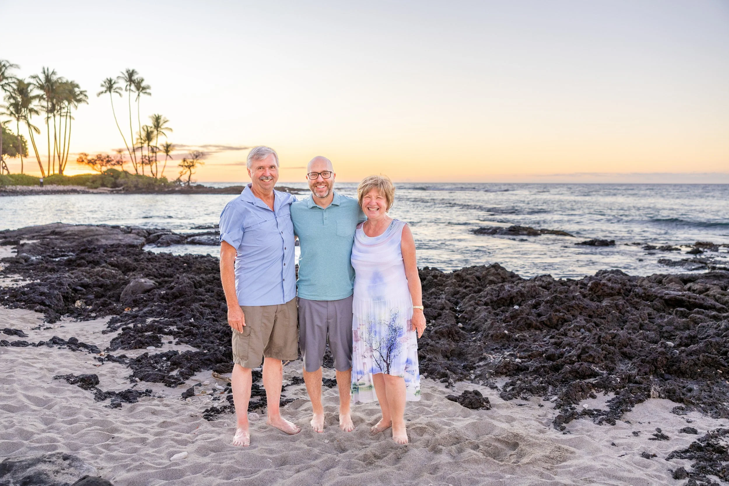 Three smiling people standing on a sandy beach with dark rocks, ocean waves, and palm trees in the background at sunset.
