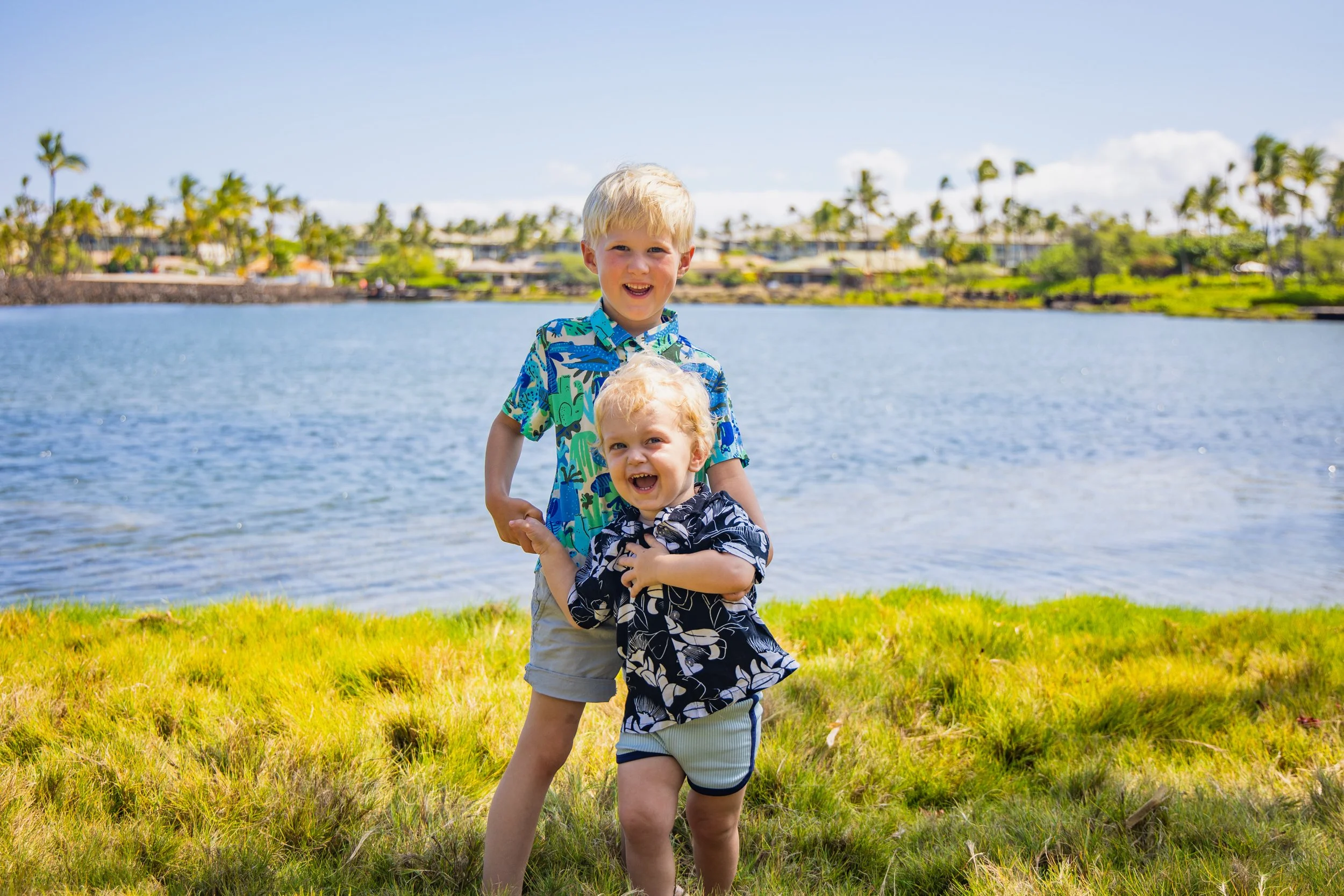 Two young boys with blonde hair smiling and playing by a lake on a sunny day, wearing colorful shirts and shorts, with palm trees and buildings in the background.