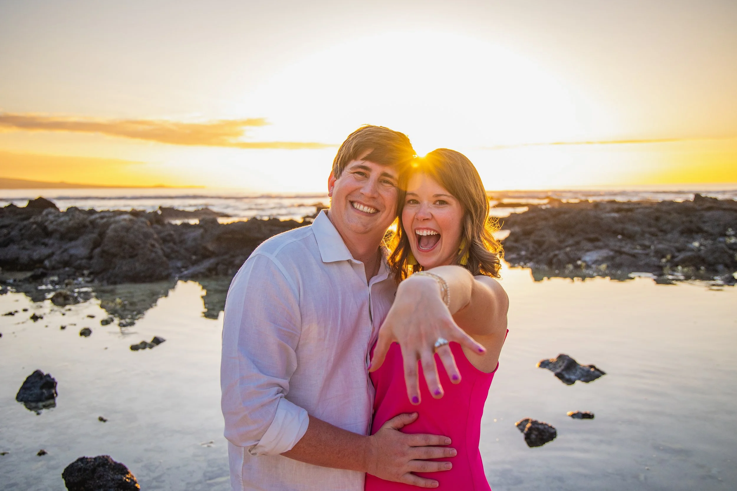 Happy couple at the beach during sunset, showing off a ring on the woman's finger.