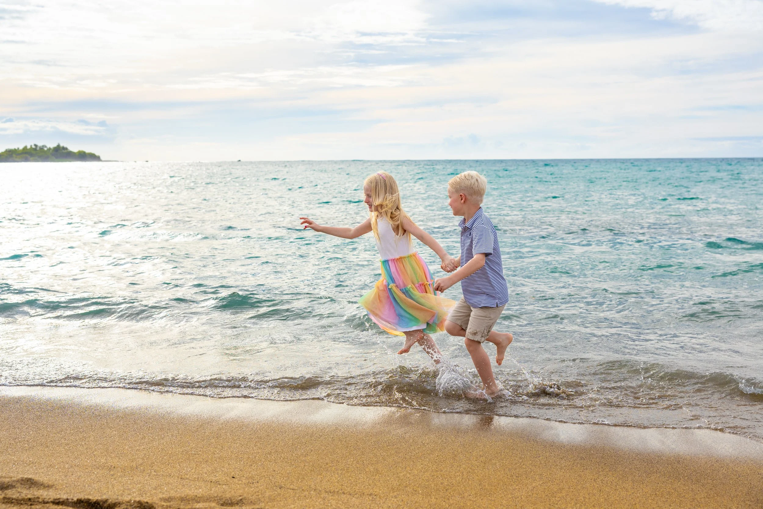 Two children, a girl in a rainbow dress and a boy in a blue striped shirt and tan shorts, playing and holding hands while running at the beach near the water.