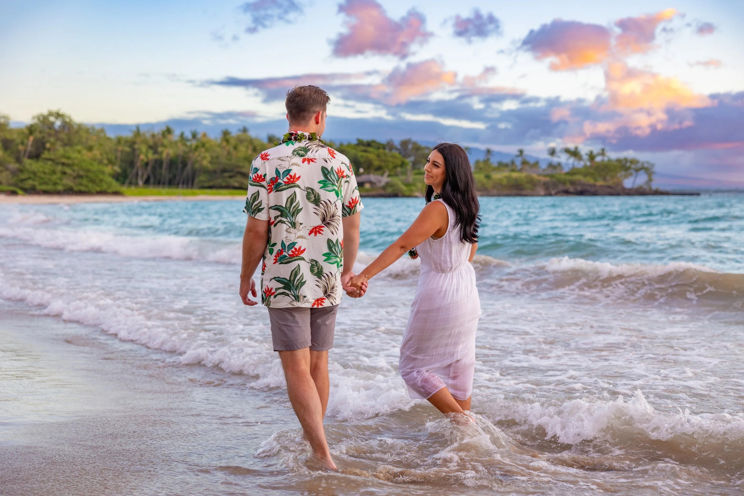 A couple holding hands and walking into the ocean at sunset, with tropical trees and a colorful sky in the background.