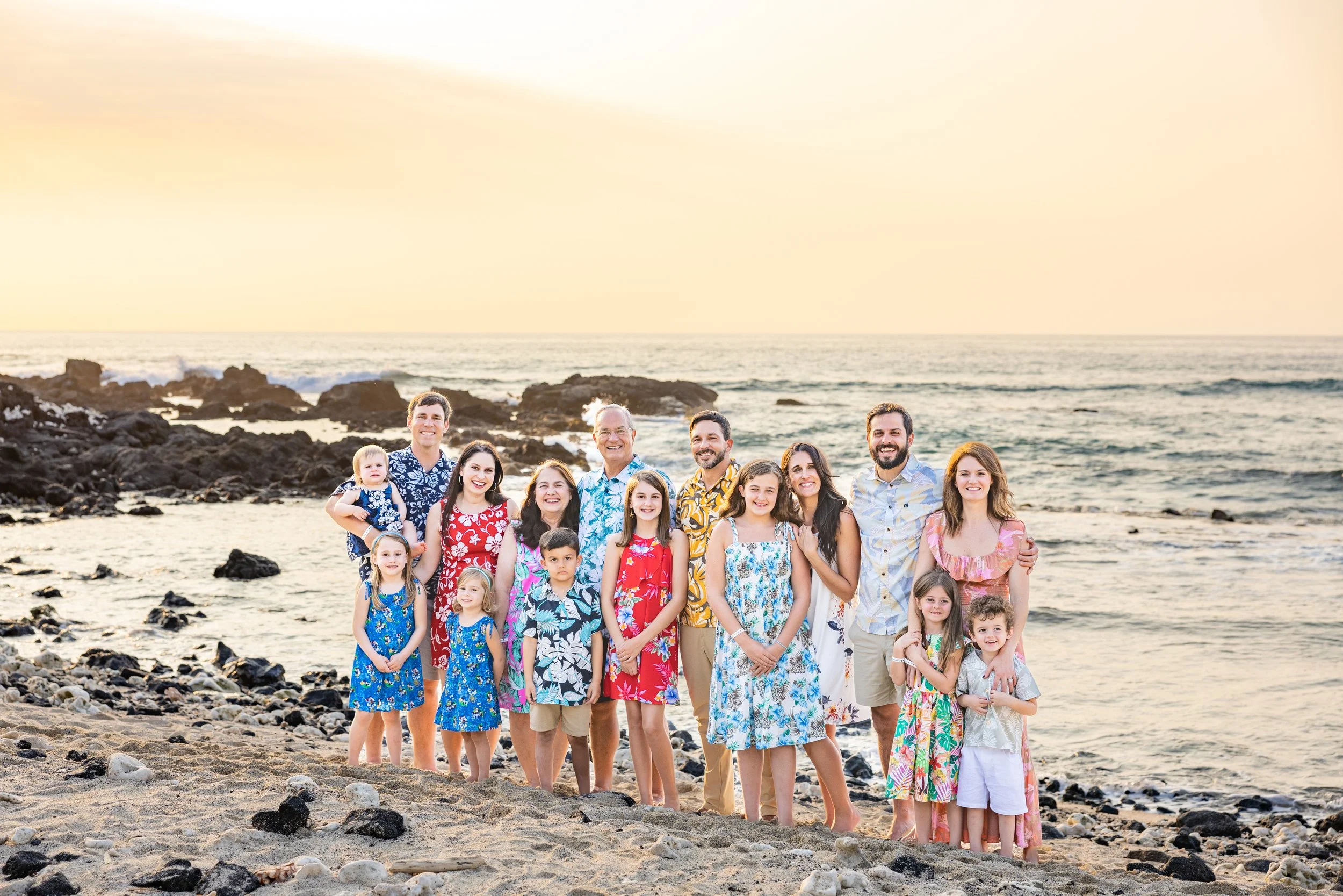 A large multigenerational family on the beach at sunset, posing for a group photo with the ocean and rocks in the background.