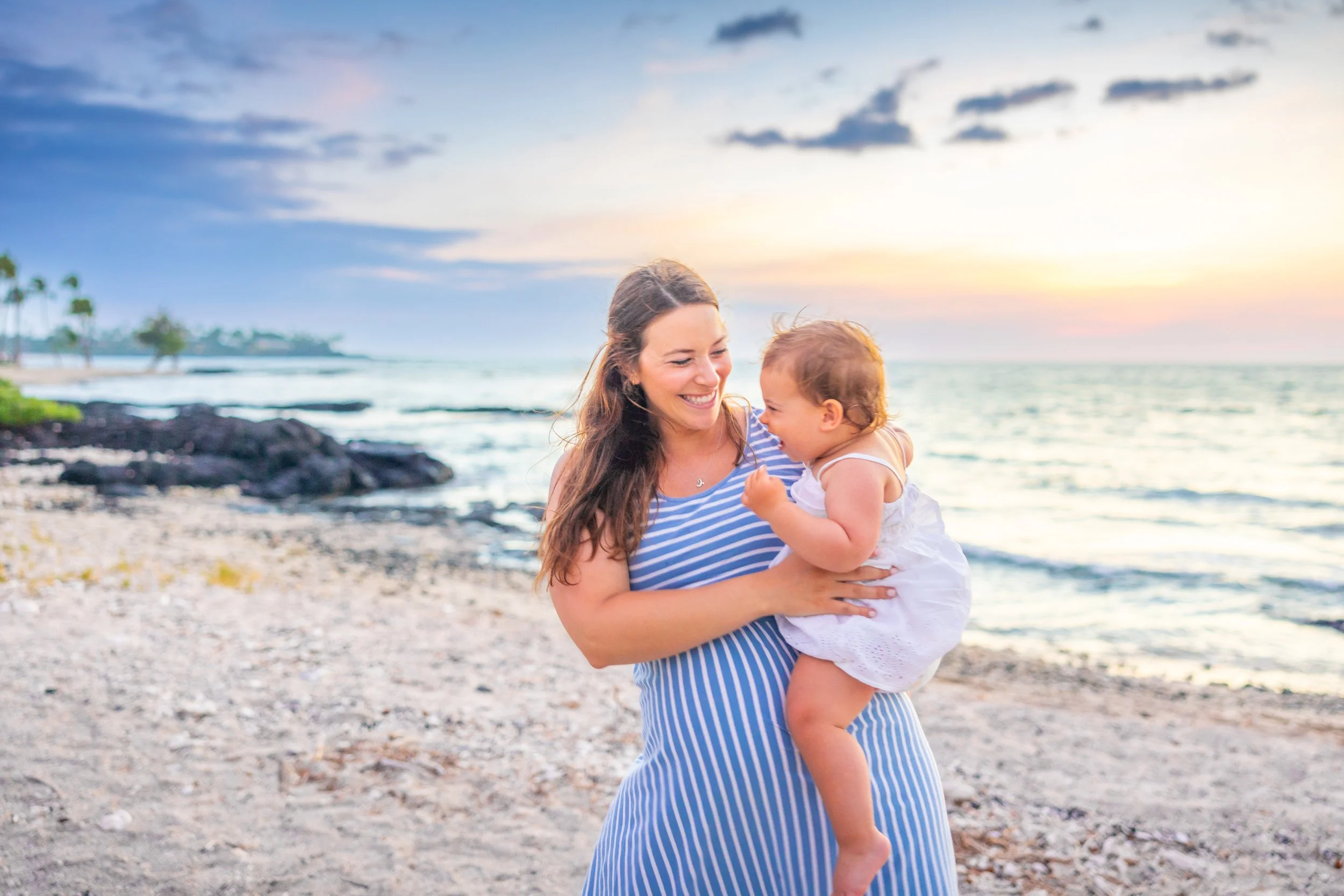 A woman holding a young girl on a beach during sunset, both smiling and enjoying each other's company.
