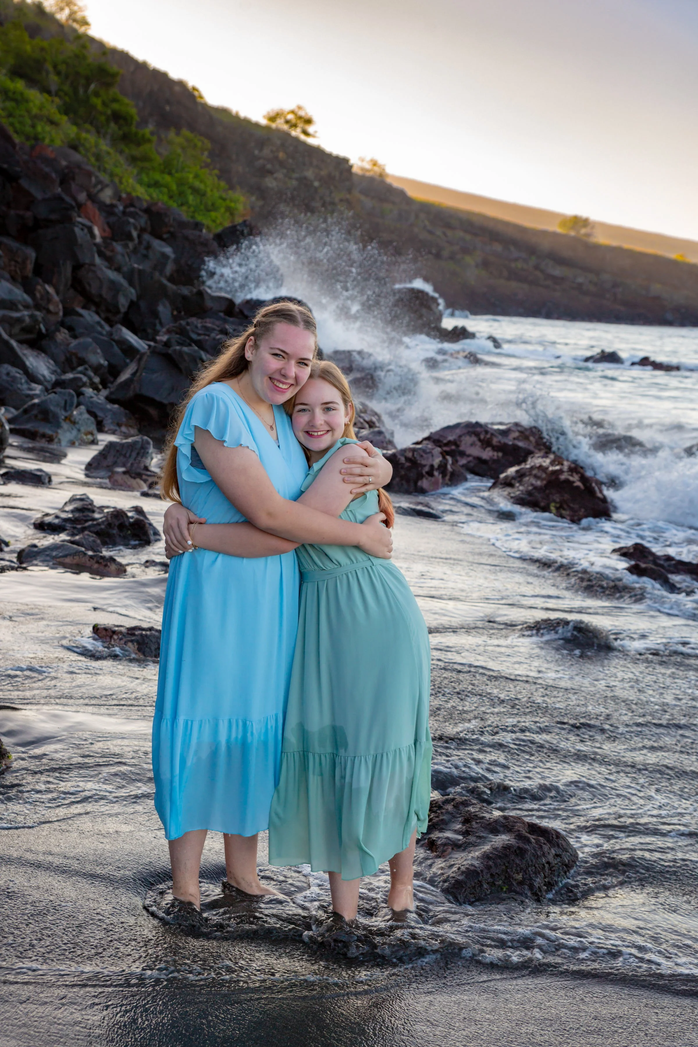 Two young women in blue dresses hugging and smiling on a rocky beach with waves crashing in the background during sunset.