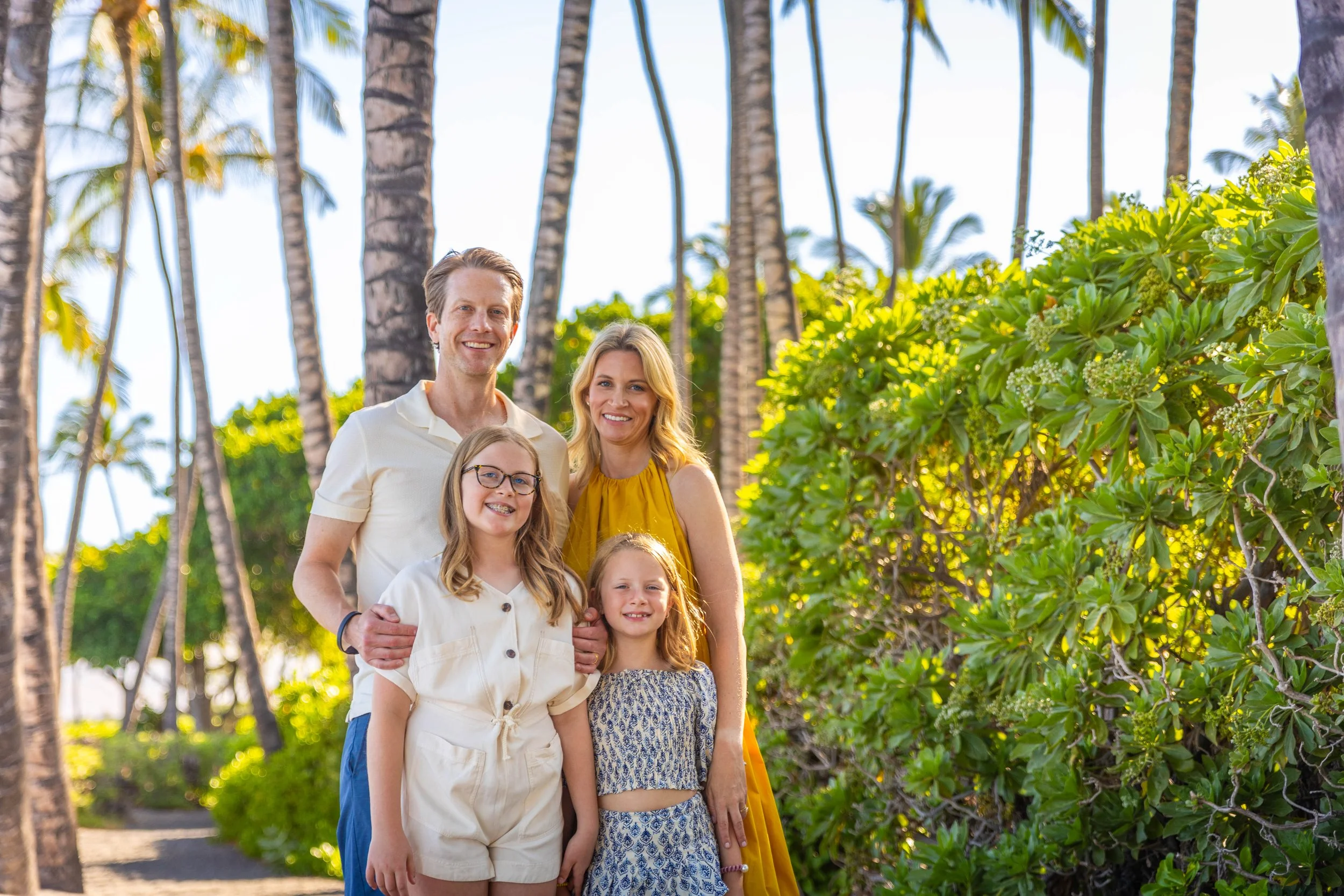 A family of four smiling and posing together outdoors in a tropical setting with palm trees and green bushes.