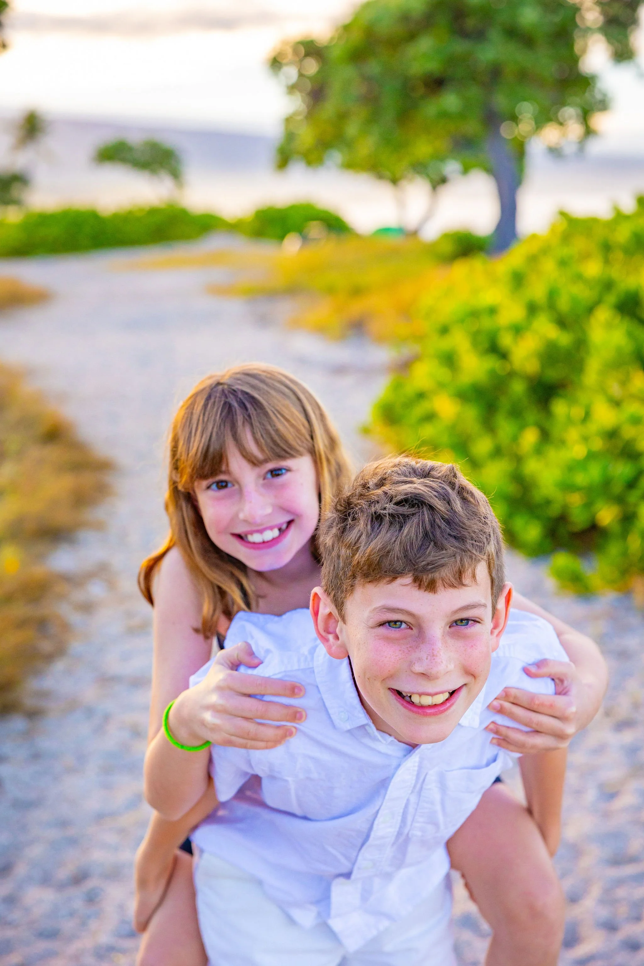Smiling children, boy giving girl a piggyback ride on a rural dirt road, trees and greenery in the background, bright and sunny weather.