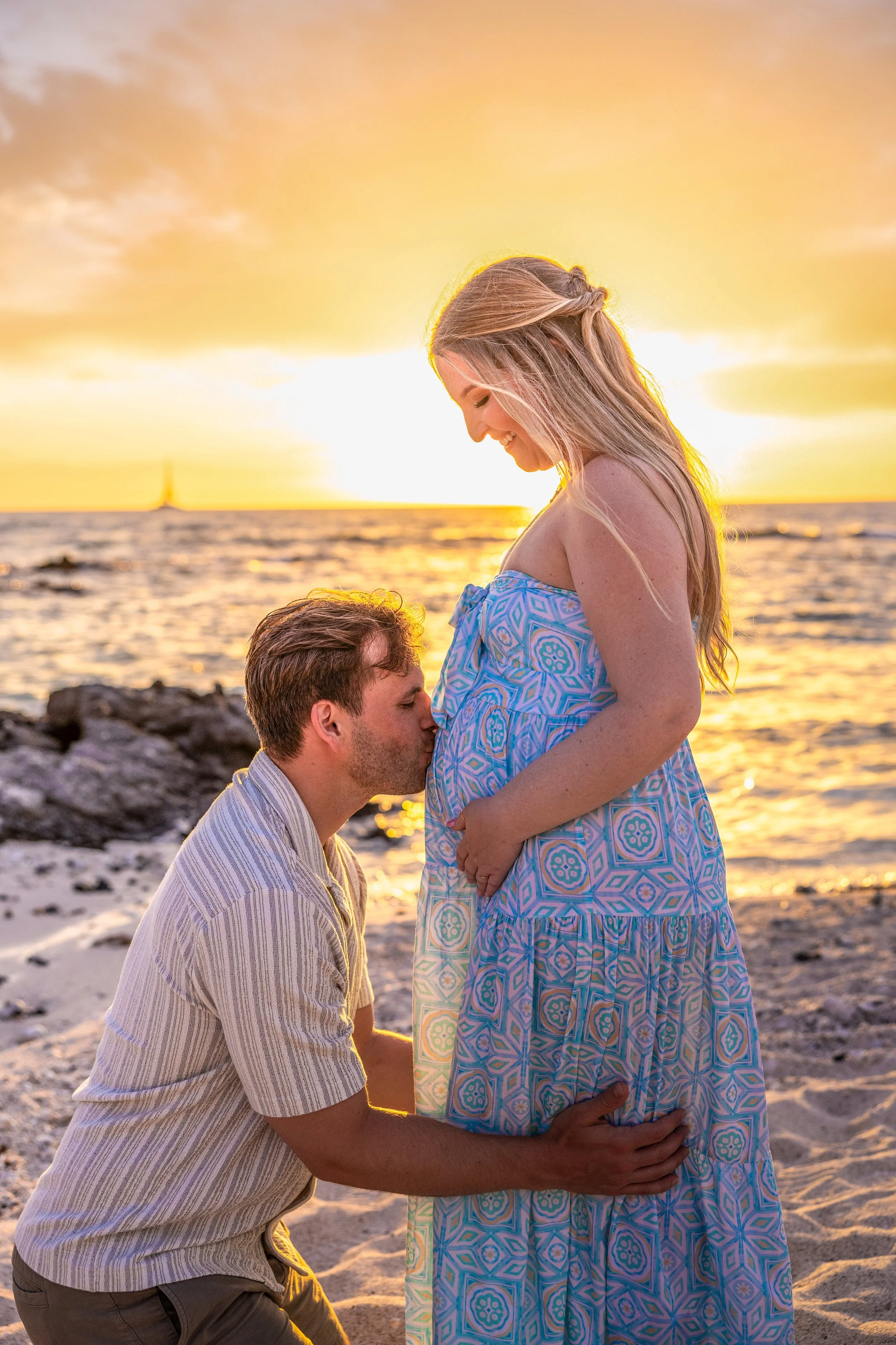 A man kneeling and kissing a pregnant woman on the belly at the beach during sunset.