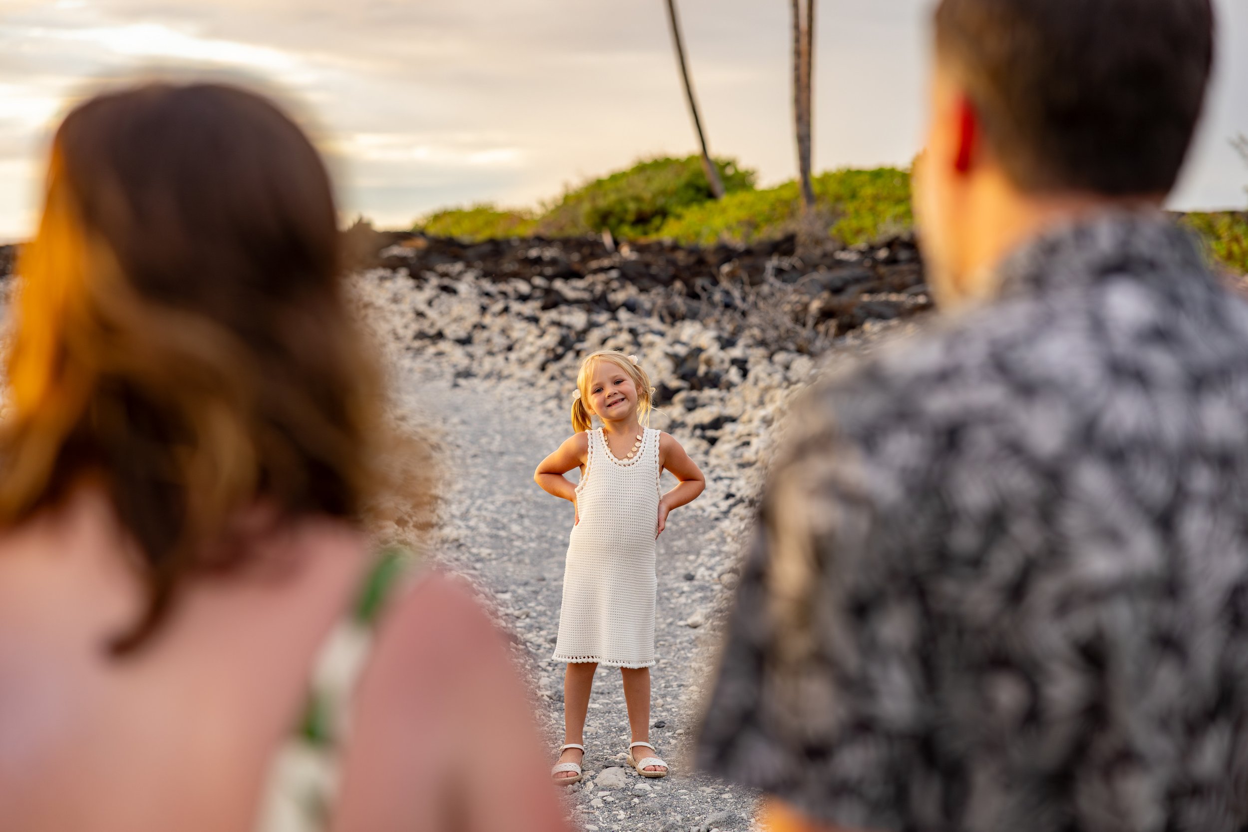 A young girl in a white dress smiling with her hands on her hips, standing on a rocky beach facing two blurred figures in the foreground, with green vegetation, rocks, and palm trees in the background during sunset.