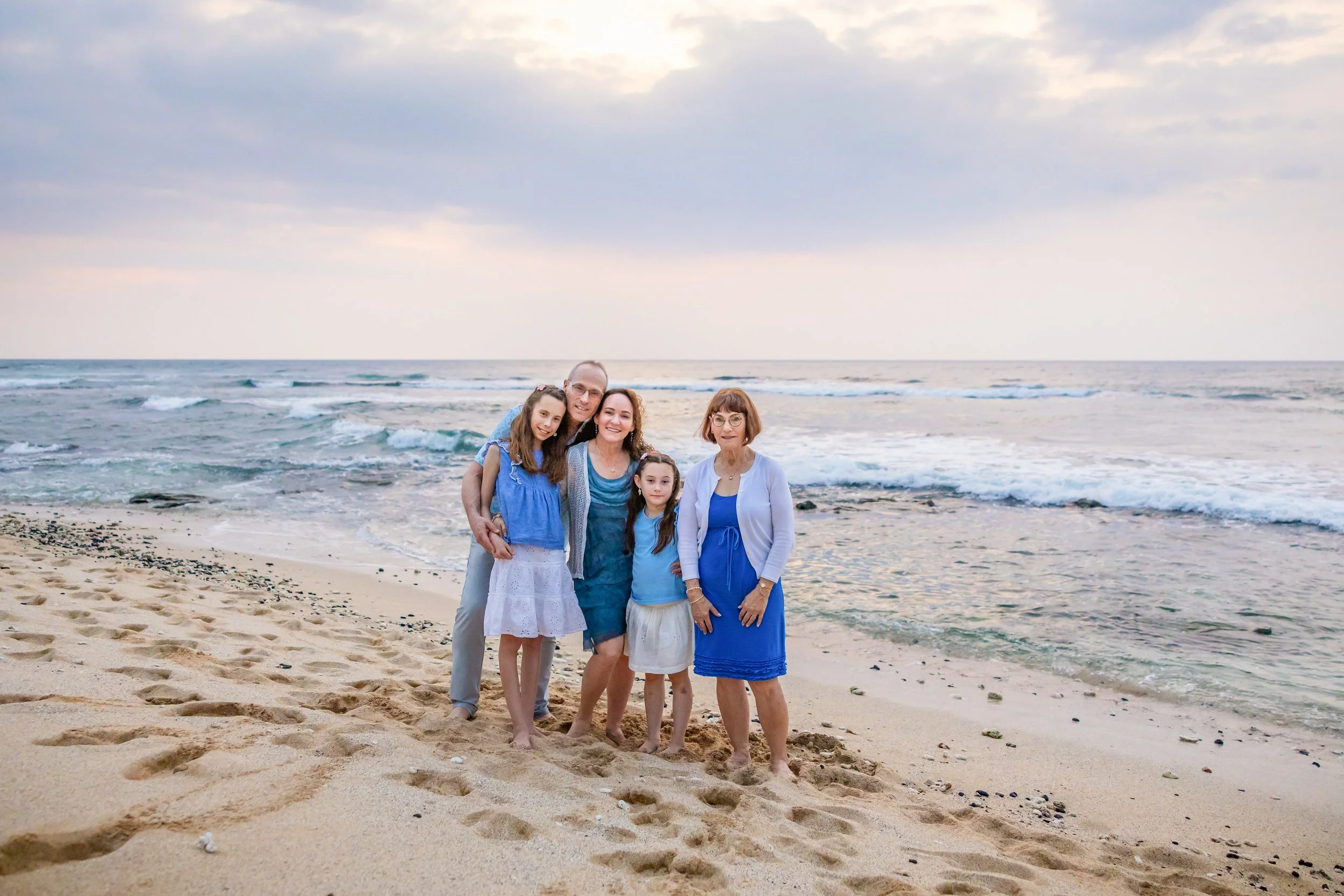 A family of five standing together on a sandy beach near the ocean at sunset.