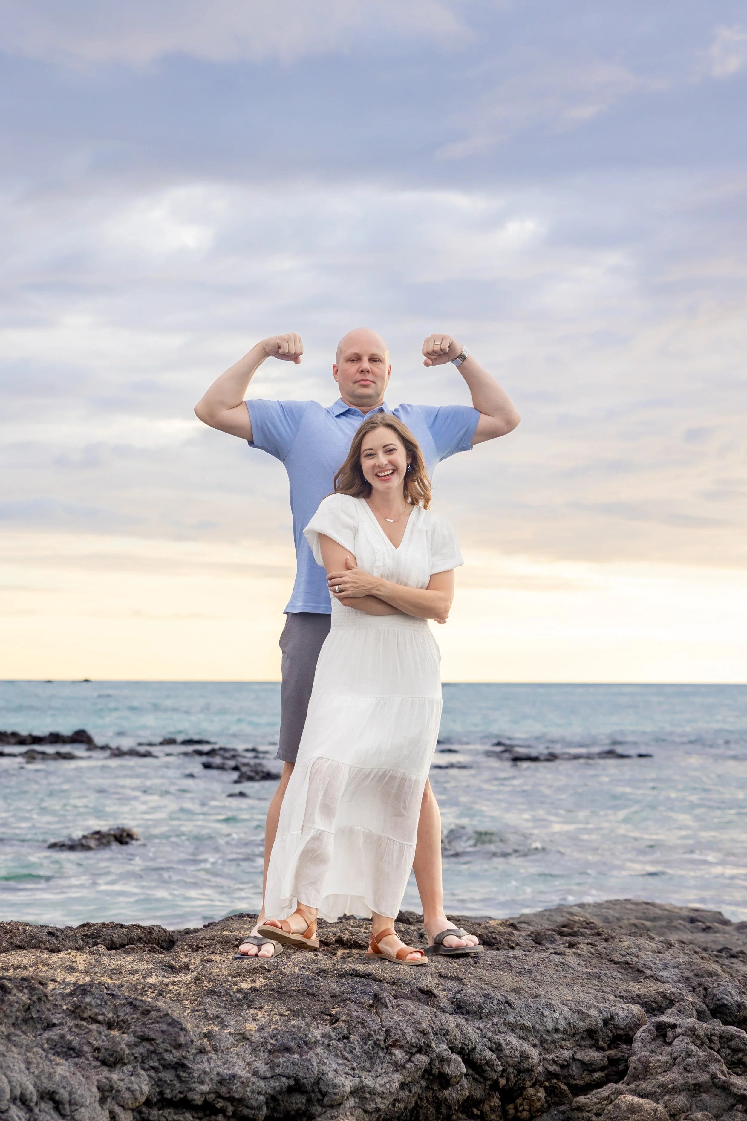 A woman standing in front with a man behind her, both on a rocky shoreline. The man is flexing his biceps while the woman is smiling. The background shows the ocean and a cloudy sky.