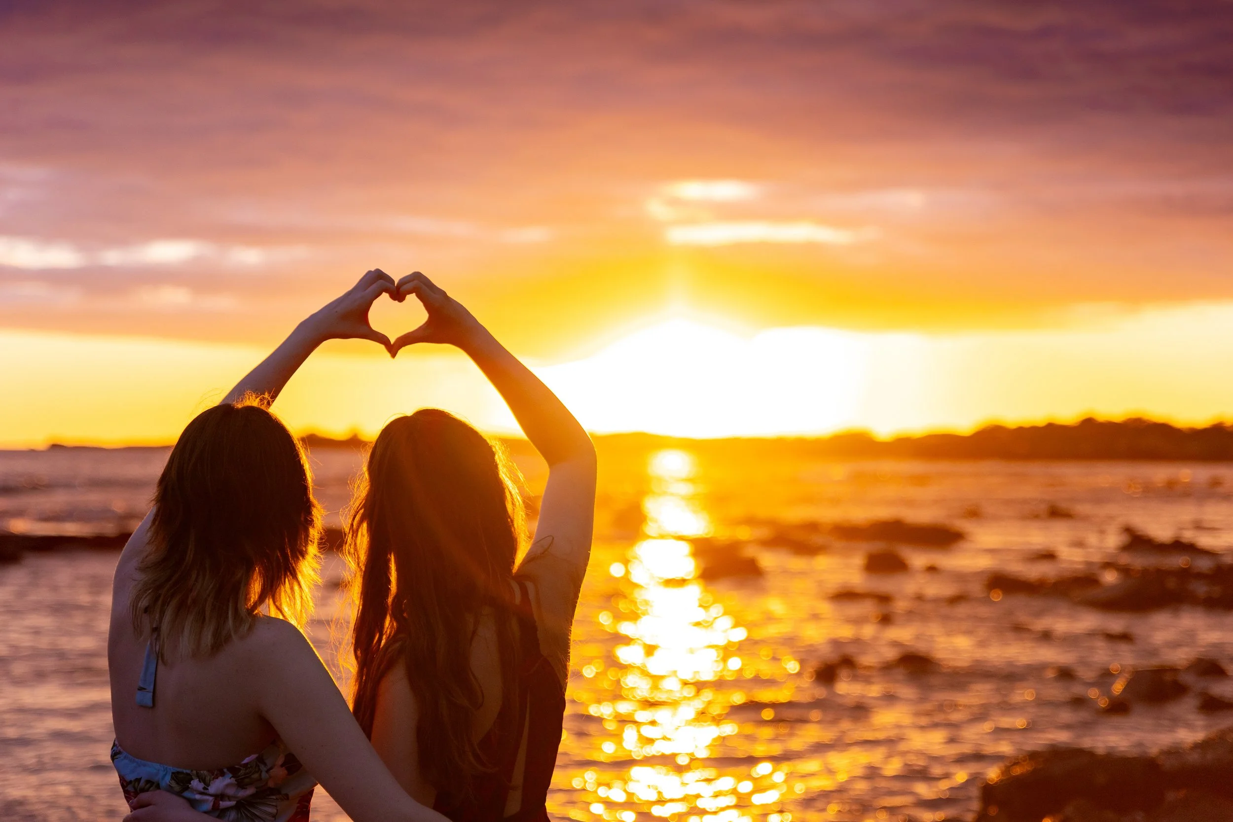 Two women at the beach during sunset, forming a heart shape with their hands against the bright sun and colorful sky.