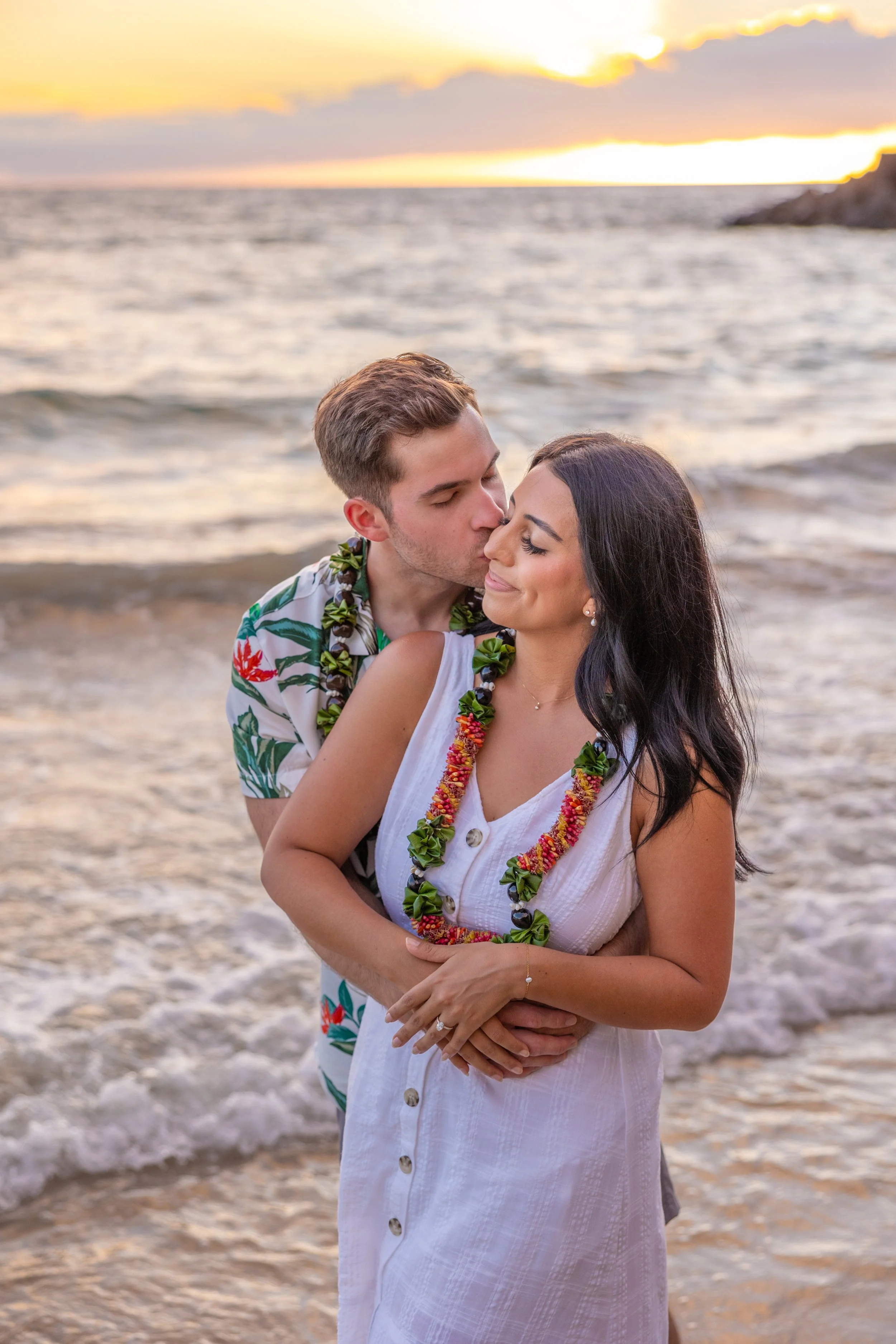 A couple stands on the beach at sunset, embracing, with the man kissing the woman's forehead. The woman wears a white dress and leis, while the man is dressed in a tropical shirt. The ocean and sunset are in the background.