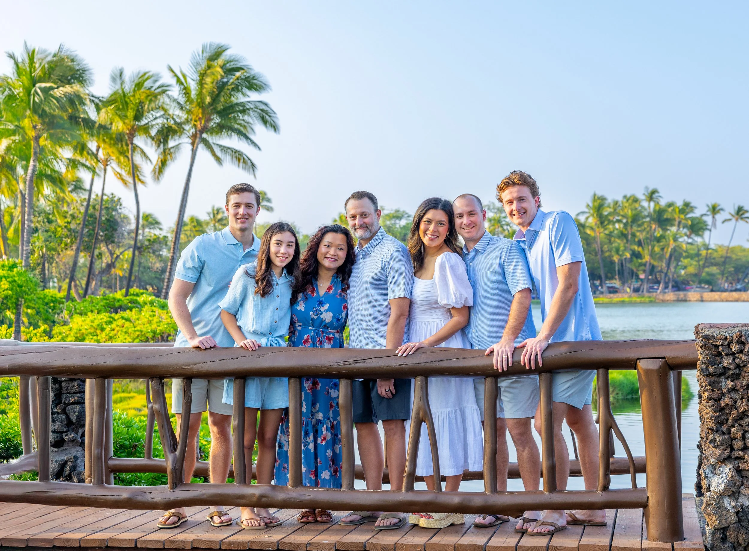 A group of seven people standing behind a wooden railing in front of a tropical landscape with tall palm trees, water, and lush greenery, smiling for the camera.”}