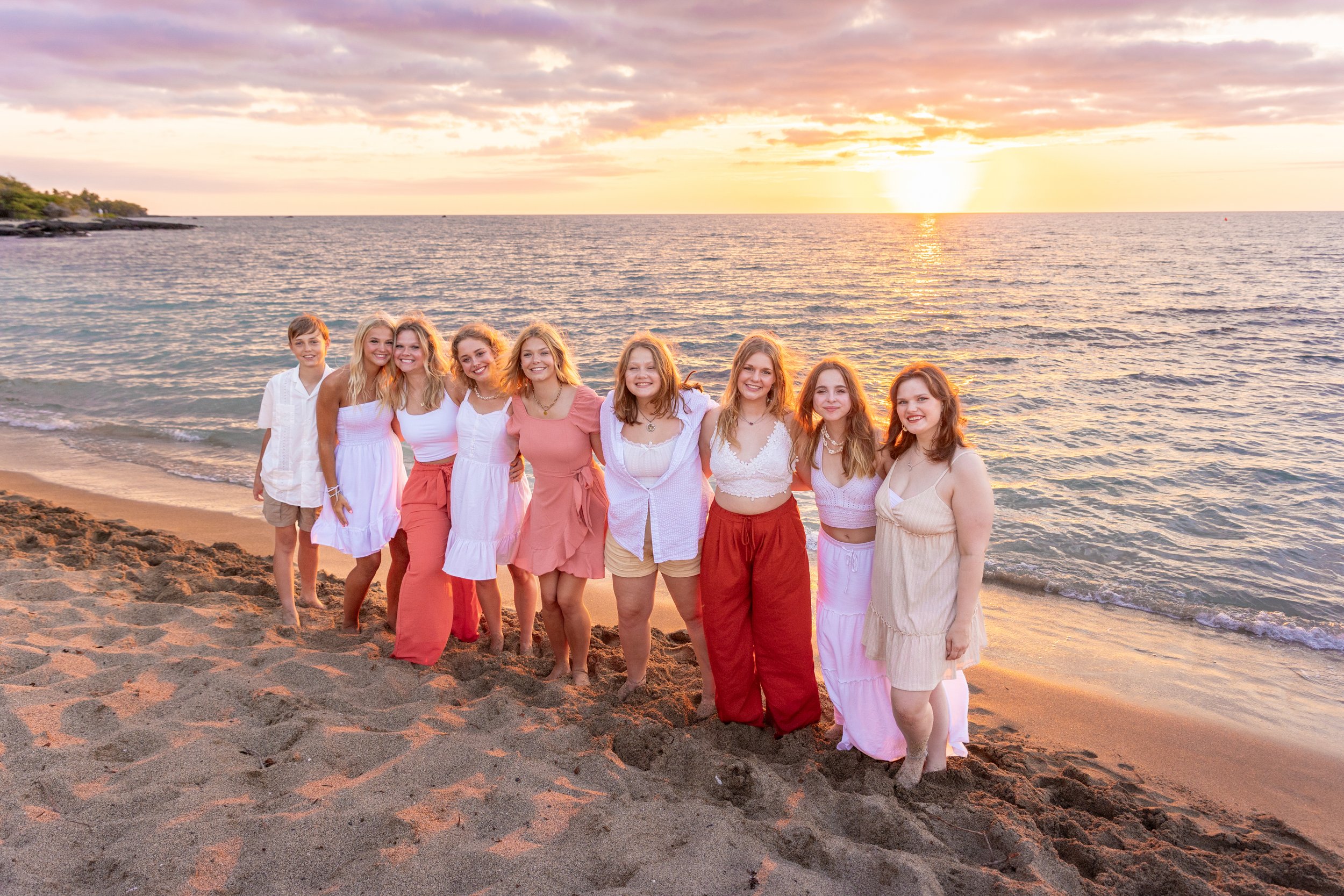 A group of ten young women and one young boy standing on a sandy beach at sunset, smiling at the camera, wearing casual summer clothing in white, pink, and beige tones with the ocean in the background.