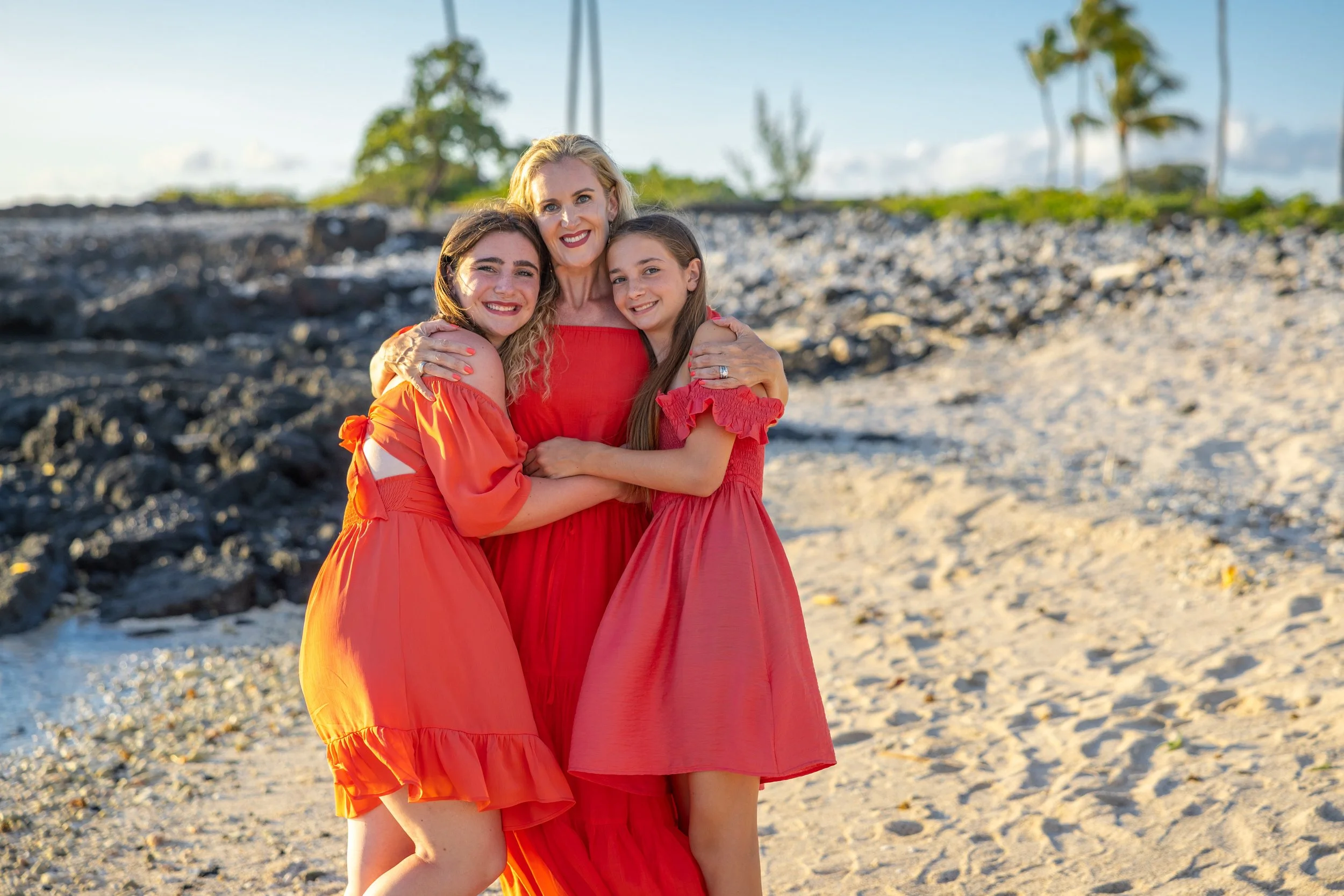Three women in red dresses hugging on a beach with sand, rocks, and palm trees in the background.