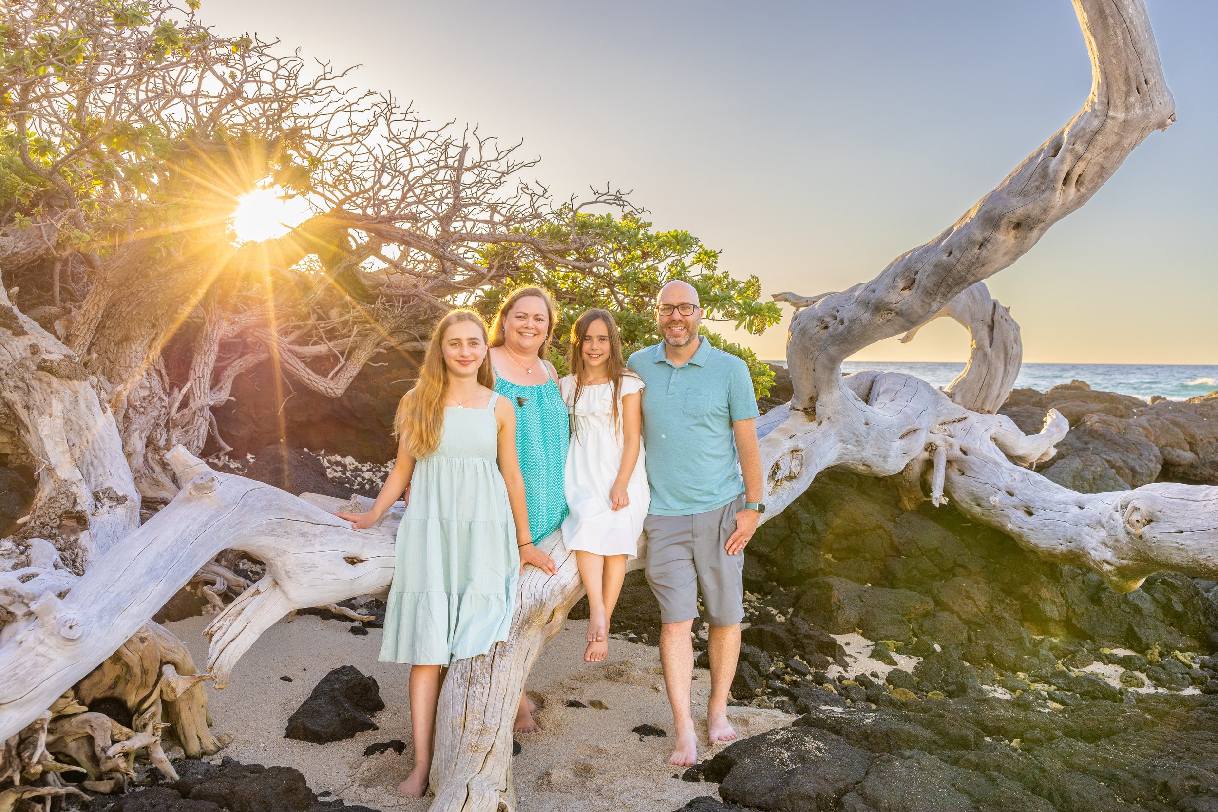Family of four standing on a beach with driftwood, rocks, and a tree, with the sun setting behind them.