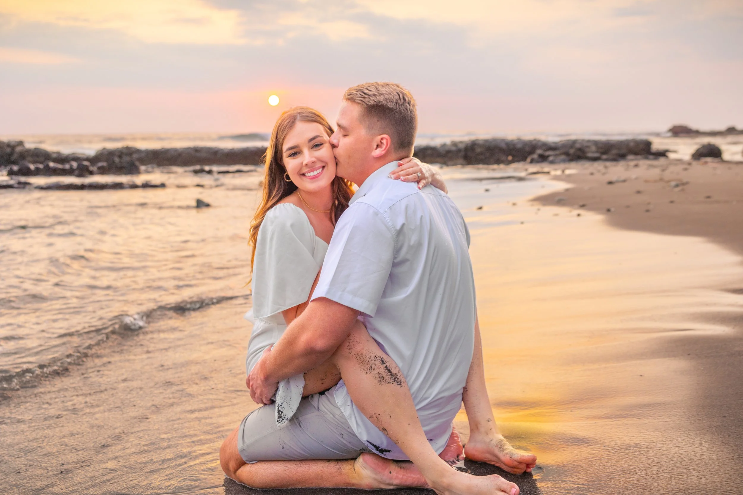 A couple sitting on beach during sunset, the woman smiling and the man kissing her cheek.