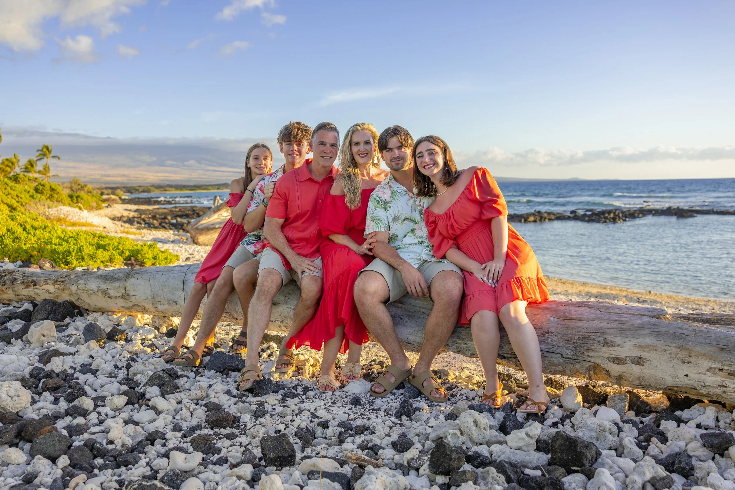 Family photo on a beach with six people sitting on a large driftwood log, wearing red and tropical print clothing, with ocean and sky in the background.