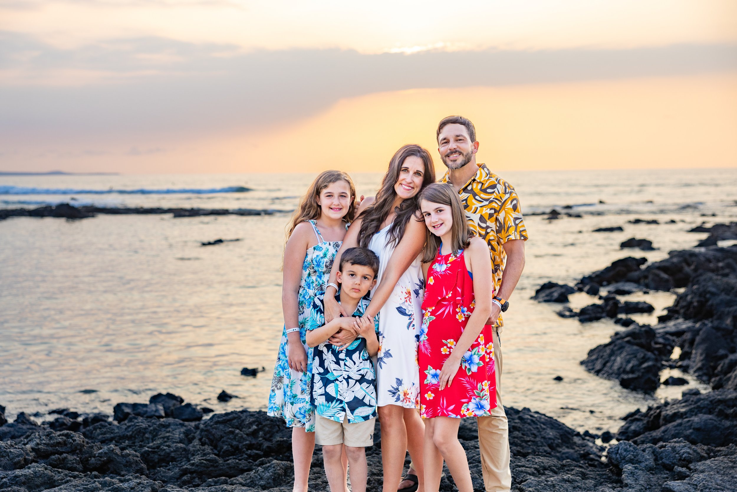 Family of six standing on rocky beach at sunset, smiling for a photo.